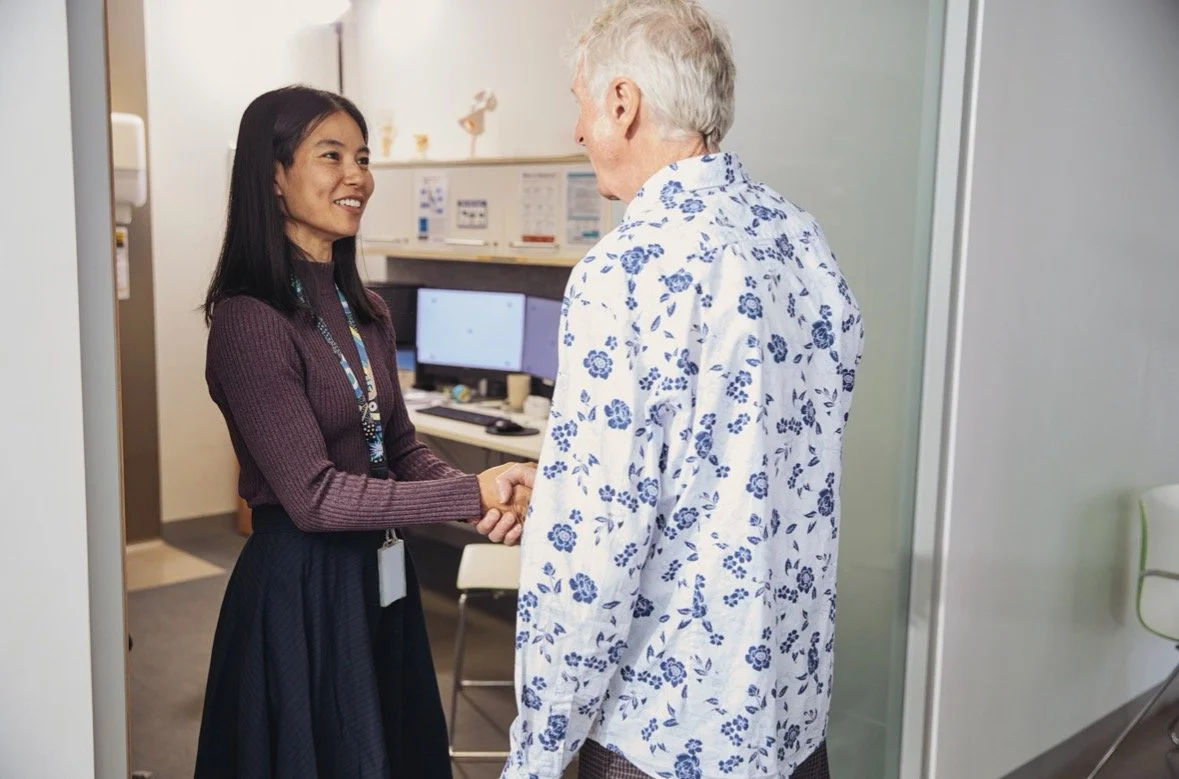 A woman and an elderly man shake hands in an office or medical setting, with computer monitors and office furniture in the background.