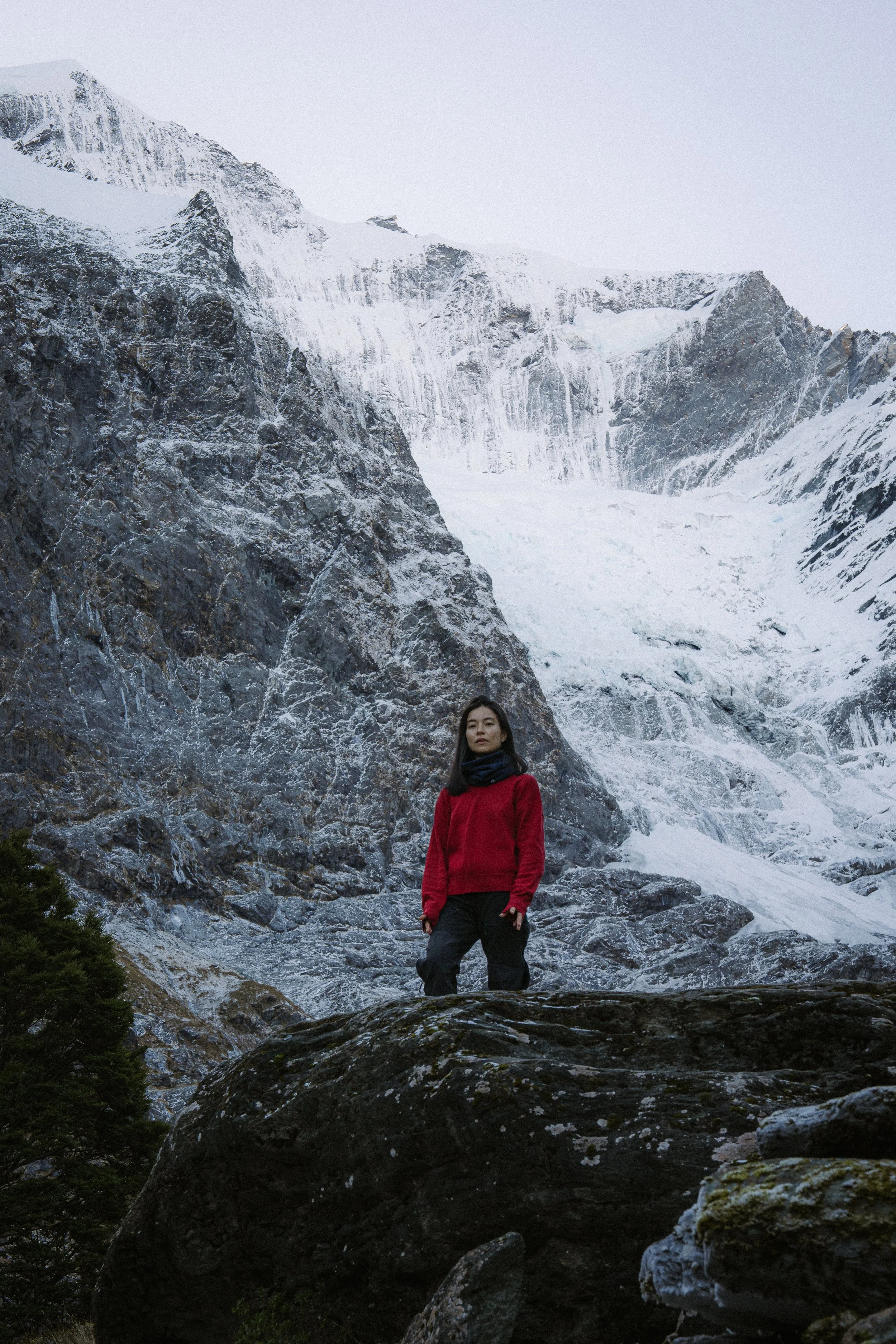 A woman in a red jacket and black pants standing on rocky terrain with snow-covered mountains in the background.