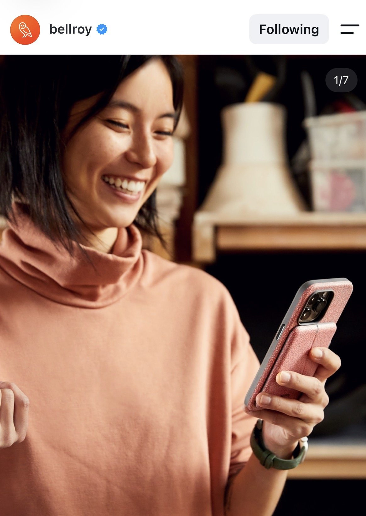 A woman with short dark hair is smiling while looking at her smartphone, which has a pink case, in a cozy indoor setting with a lamp and shelves in the background.