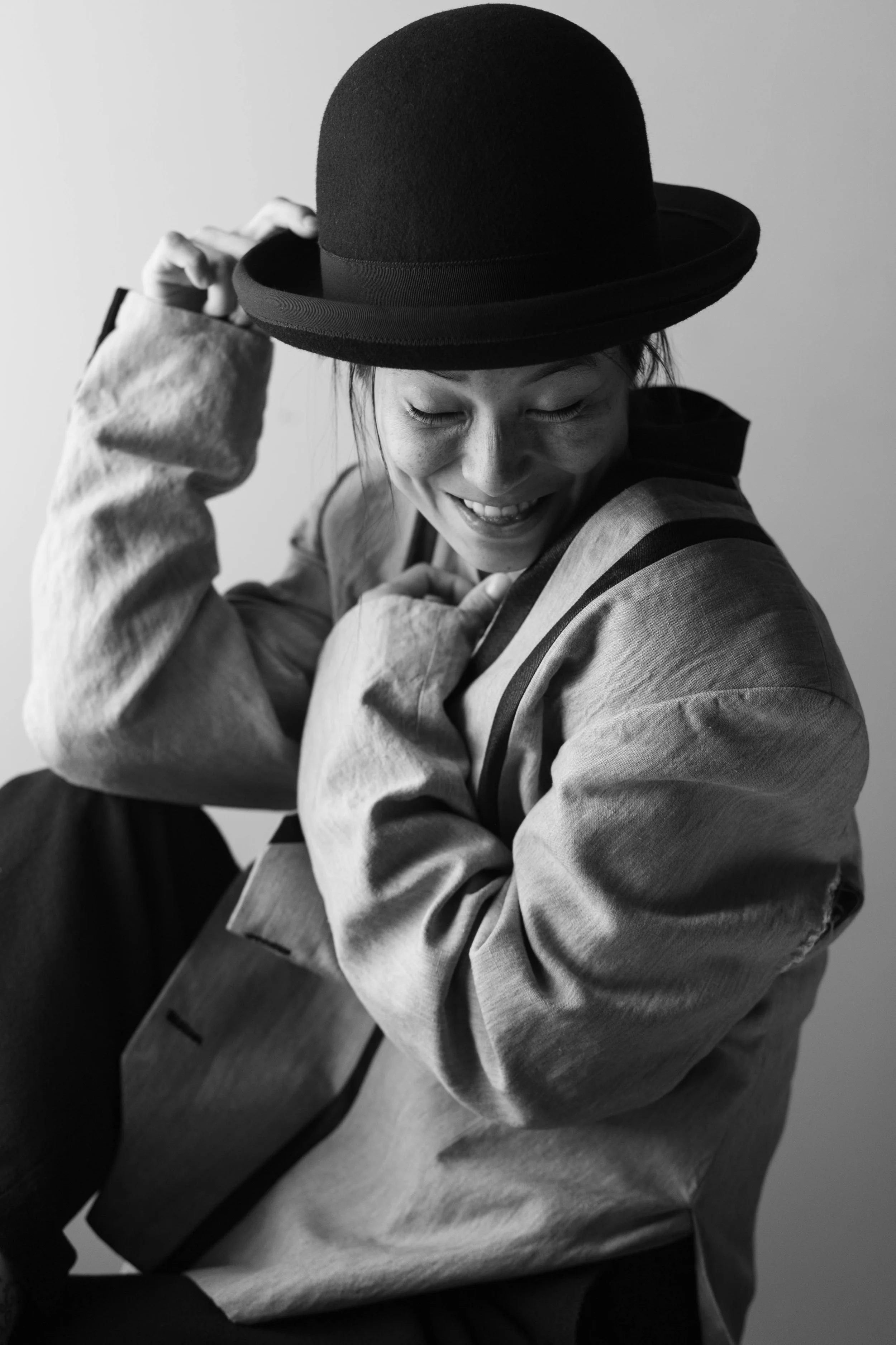 A person with short hair wearing a wide-brimmed hat and a loose gray jacket, smiling with eyes closed, sitting in a relaxed pose against a light background.