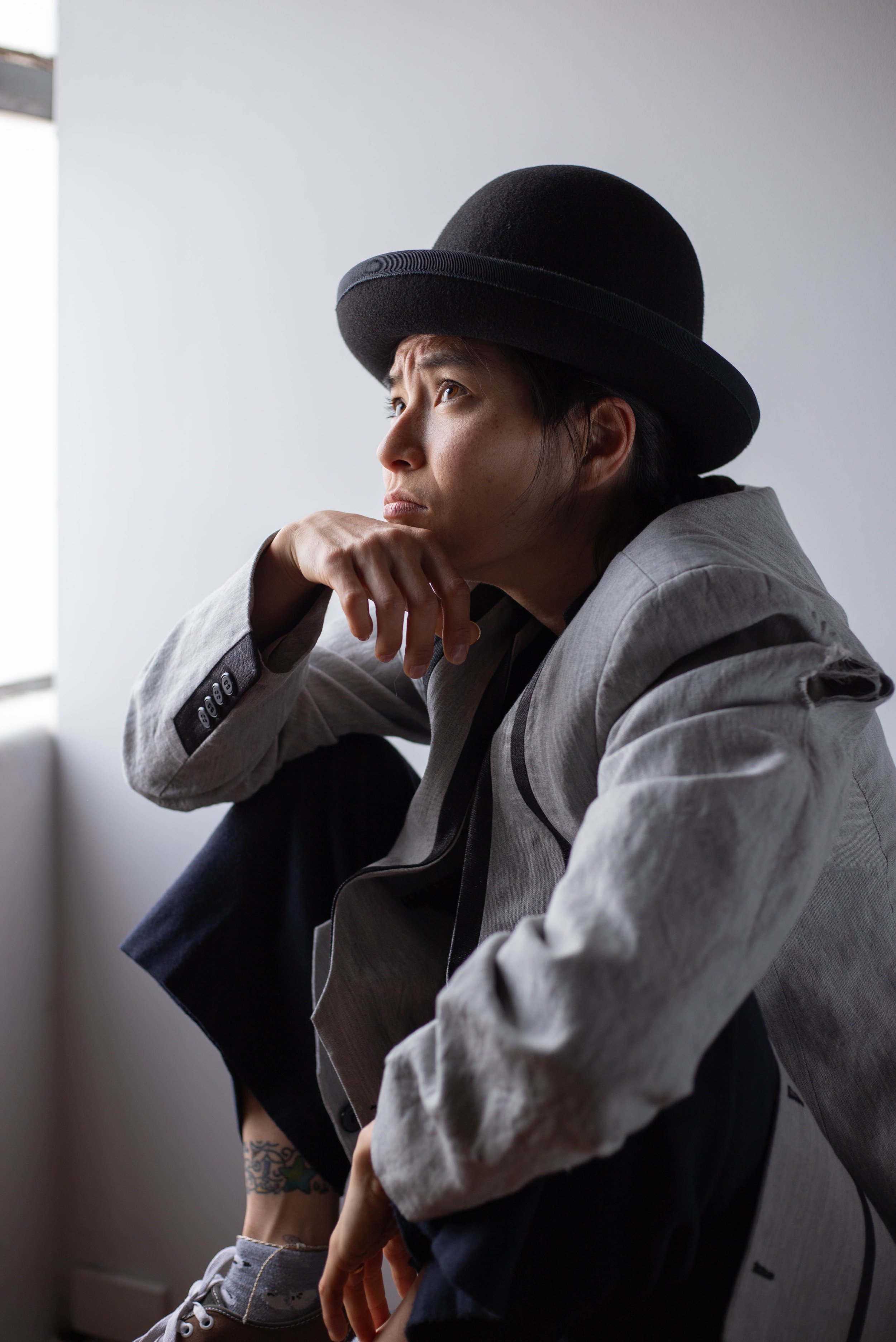A woman with short dark hair wearing a large black hat and a gray blazer, sitting indoors with her chin resting on her hand, looking contemplative. She is wearing gray sneakers and has tattoos on her ankle.