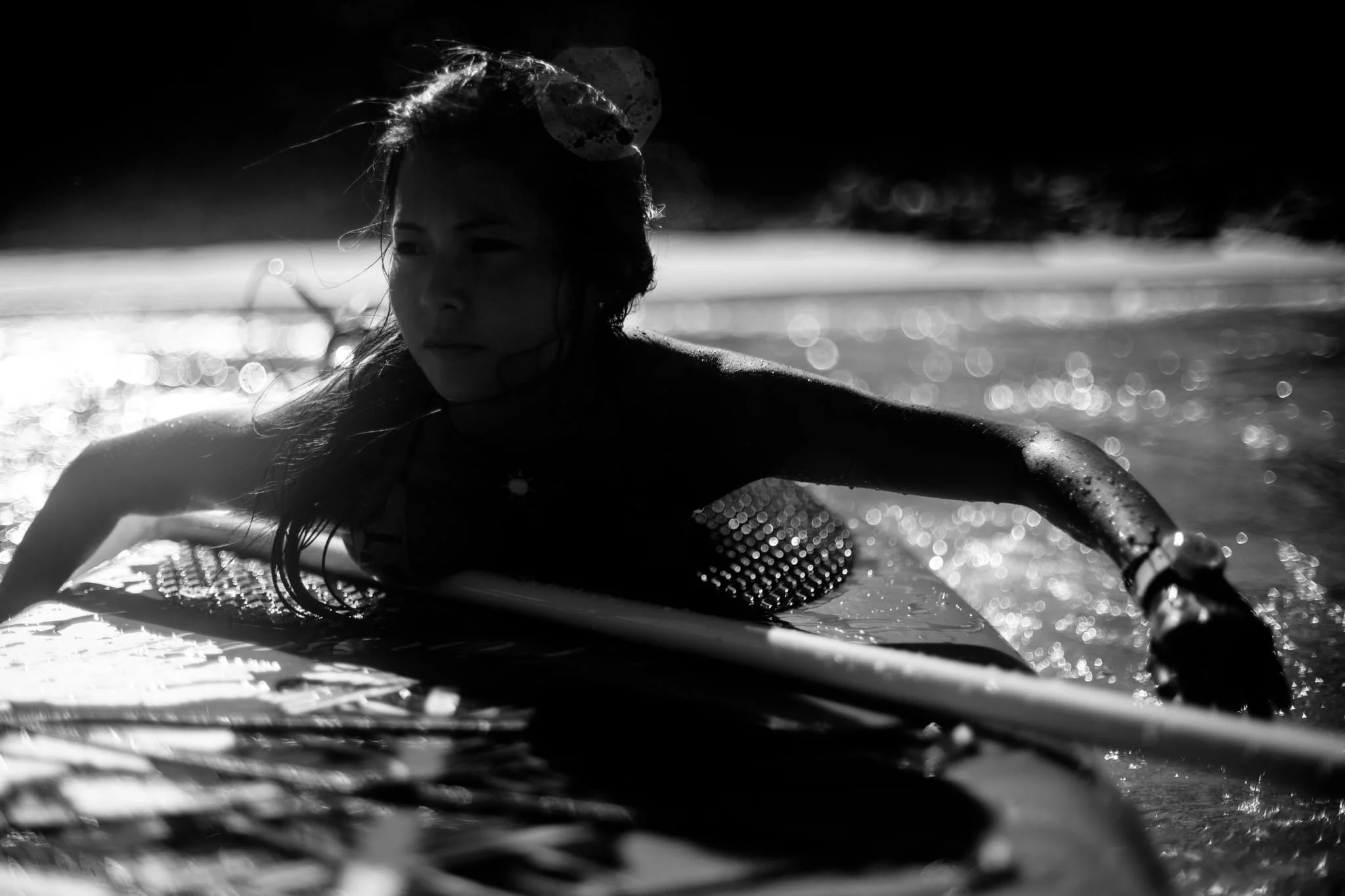 Black and white photo of a woman with wet hair in water, looking toward the camera with a serious expression, illuminated by sunlight creating bokeh effects on the water surface.