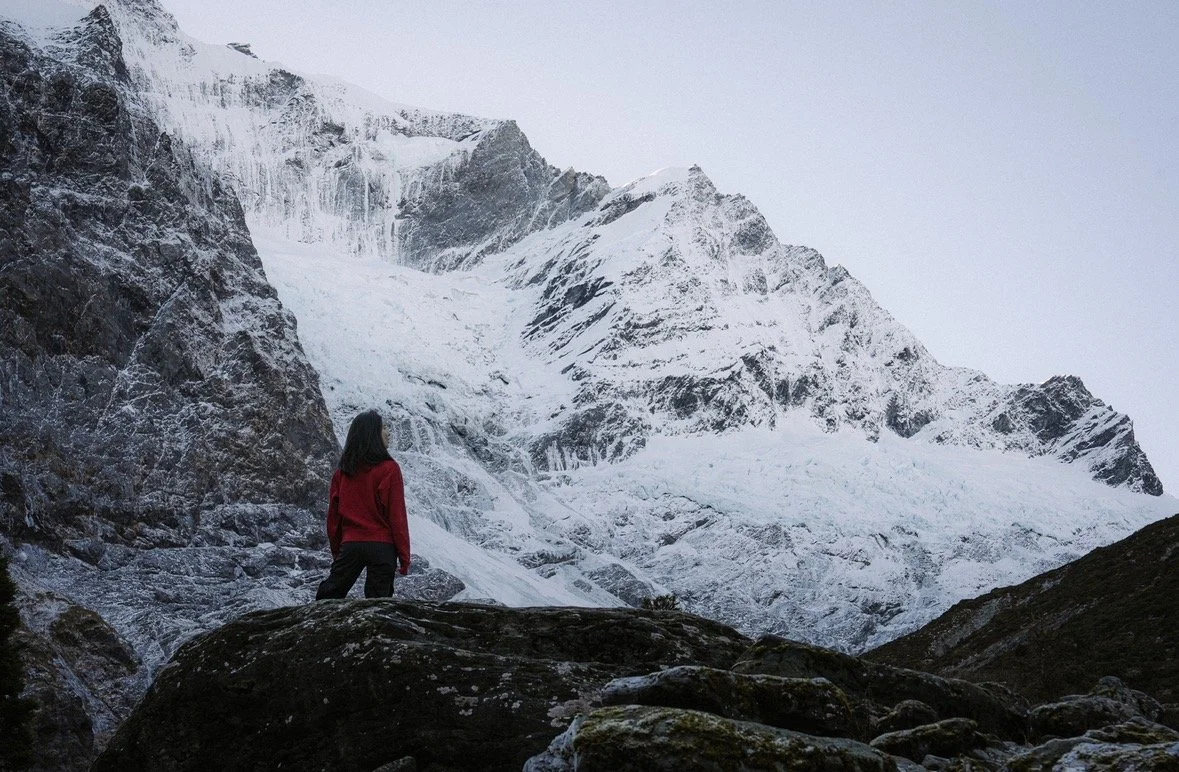 A person in a red jacket standing on rocks in front of a snowy mountain landscape with glaciers.