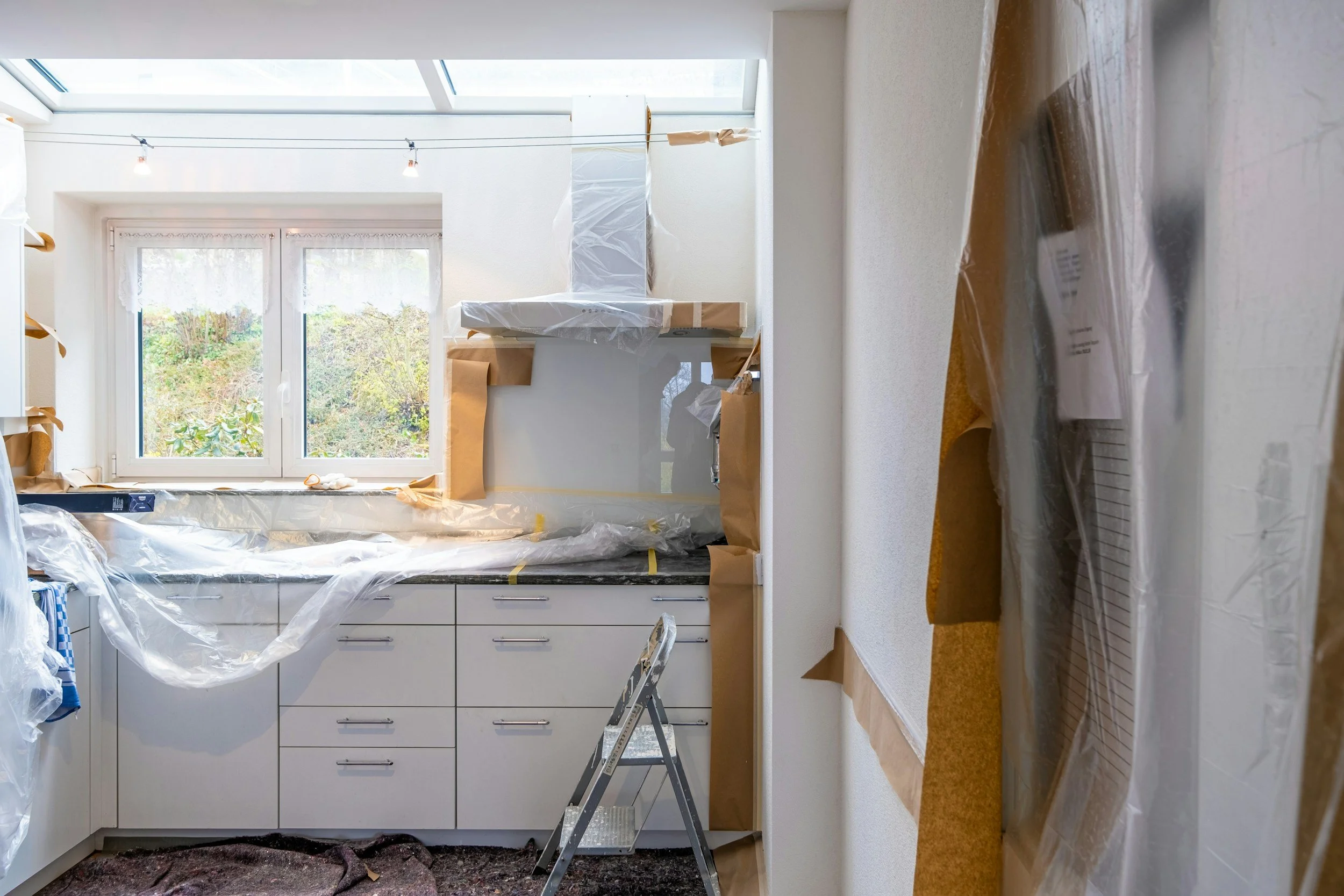 Kitchen during renovation with plastic coverings on cabinets and appliances, ladder, and a window with a view of trees outside.