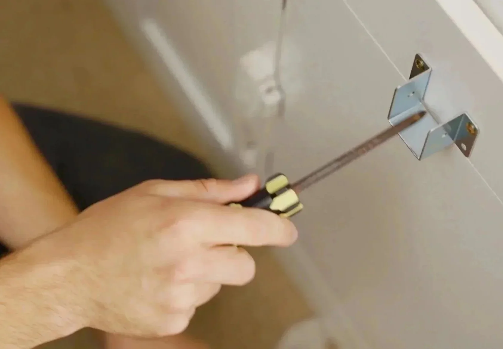 Close-up of a person's hand using a screwdriver to tighten or loosen a screw on a metal hinge attached to a door or panel.