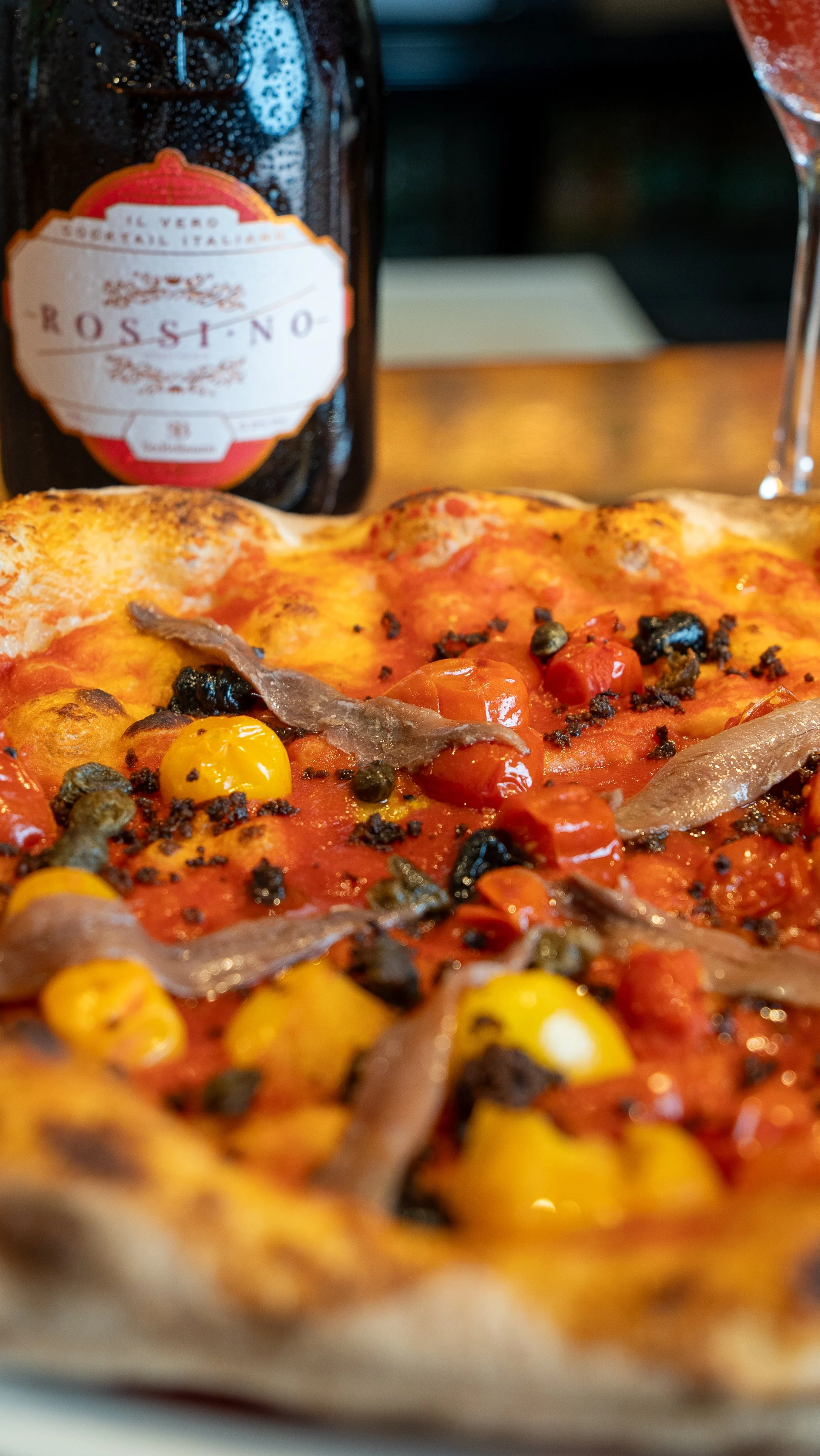 Close-up of a pizza with cherry tomatoes, black olives, anchovies, and yellow peppers, with a bottle of Rosso wine behind it.