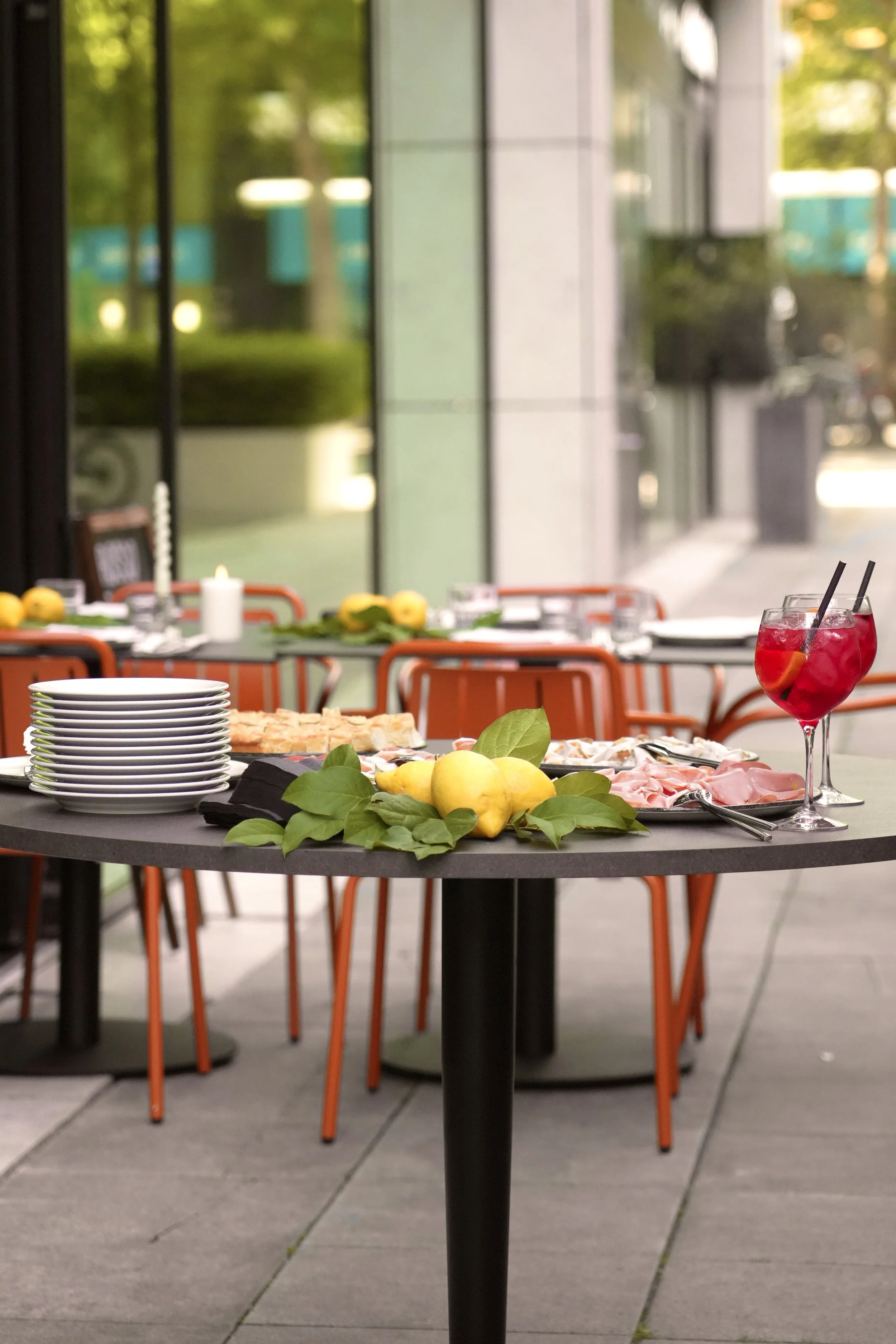 A table set for a meal with plates, lemons, and a red drink with ice and two straws, in front of large glass windows and orange chairs outside.