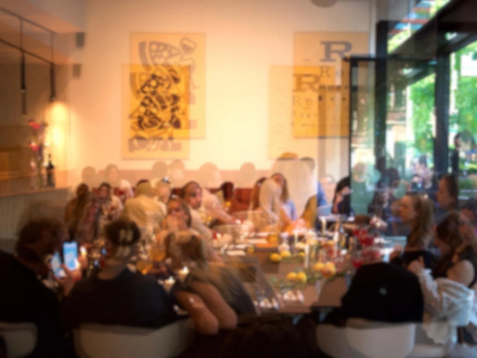 A lively indoor gathering with people seated at tables during daytime, some of whom are taking photos. The room has large windows showing green trees outside, and artwork hangs on the walls.