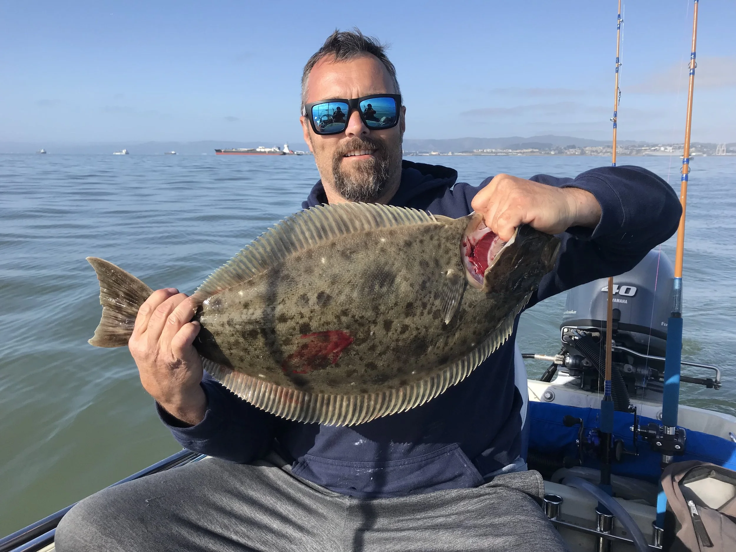 A man with sunglasses and a beard sitting in a boat, holding a large fish he caught, with boats and a shoreline visible in the background.