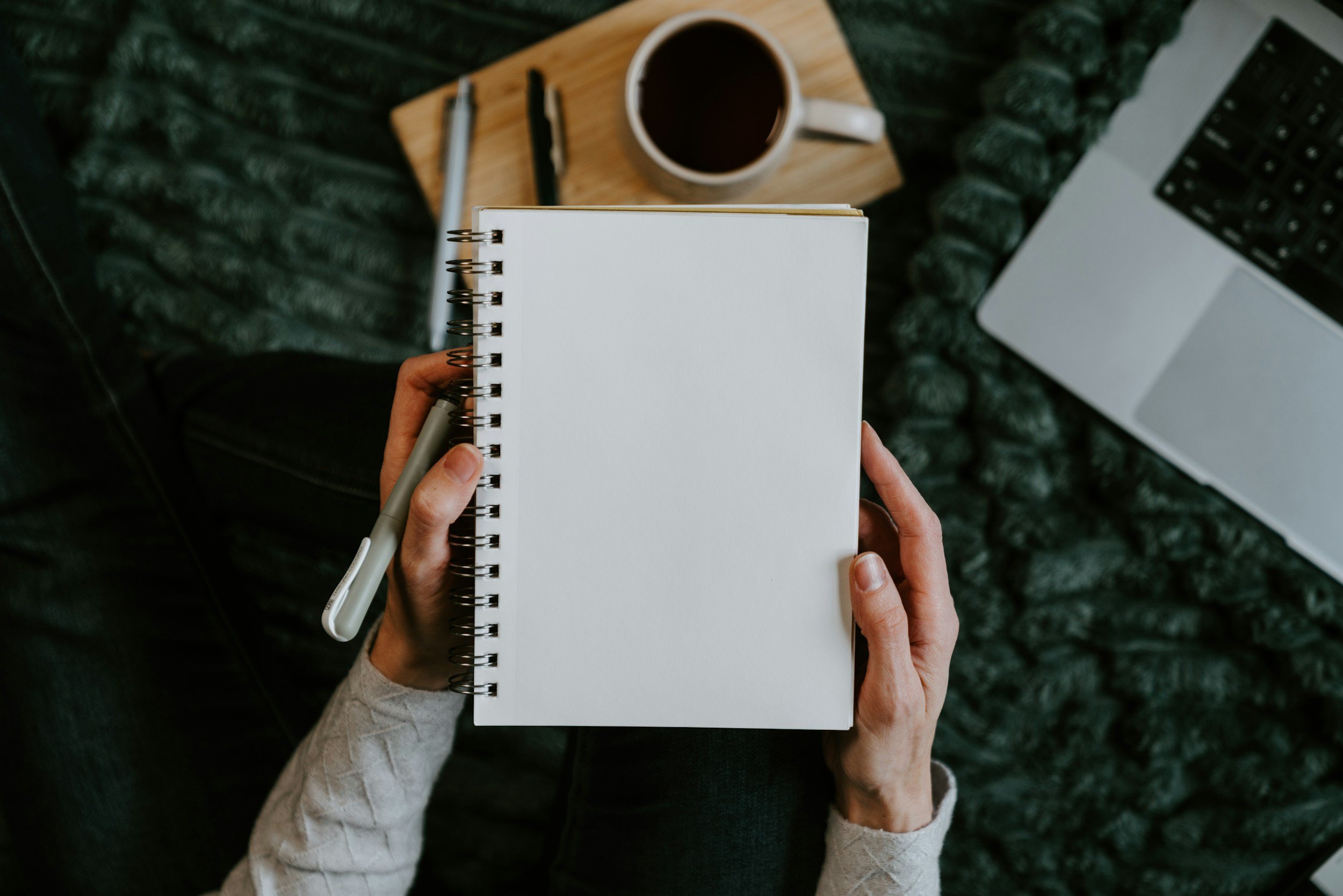 Person holding a notebook and pen while sitting at a desk with a cup of coffee, a keyboard, and a laptop.