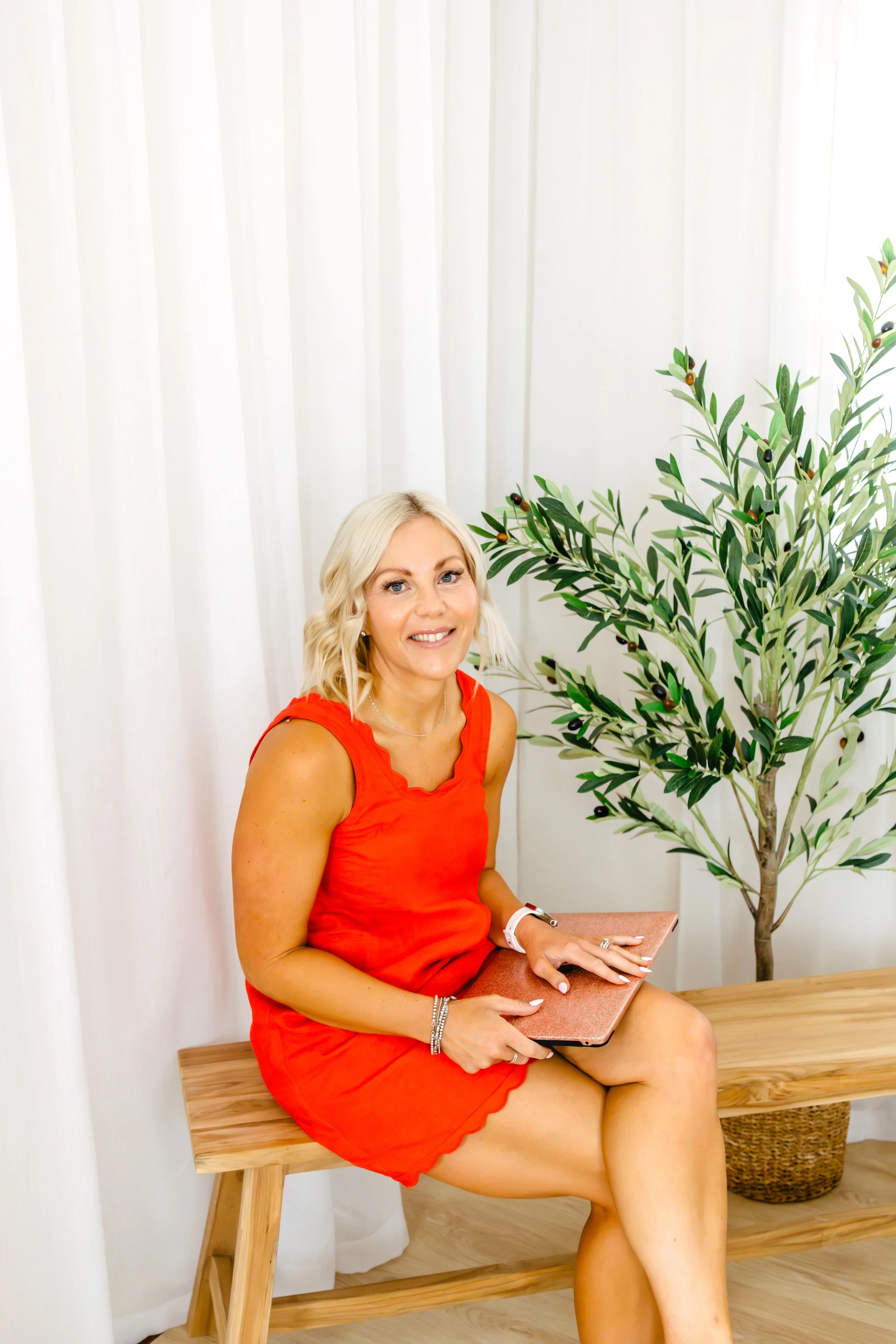 A smiling blonde woman in a red dress sitting on a wooden bench with a tablet in her lap, next to a potted plant with green leaves and black berries, in a room with white curtains.