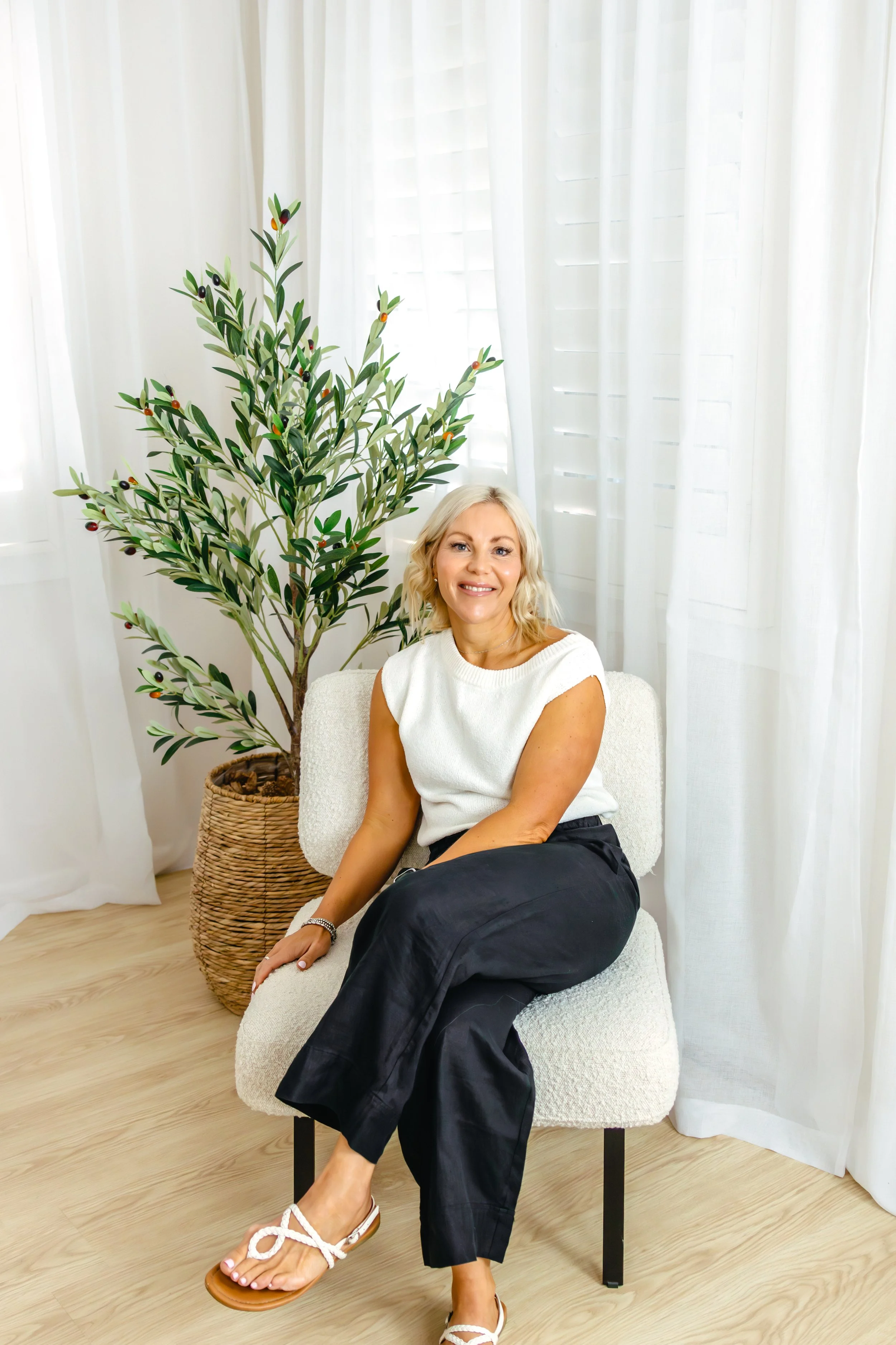 A woman with blonde hair in a white sleeveless top and black pants sitting on a white chair, smiling, with a large olive tree in a basket behind her, white curtains in the background, and light-colored wooden flooring.