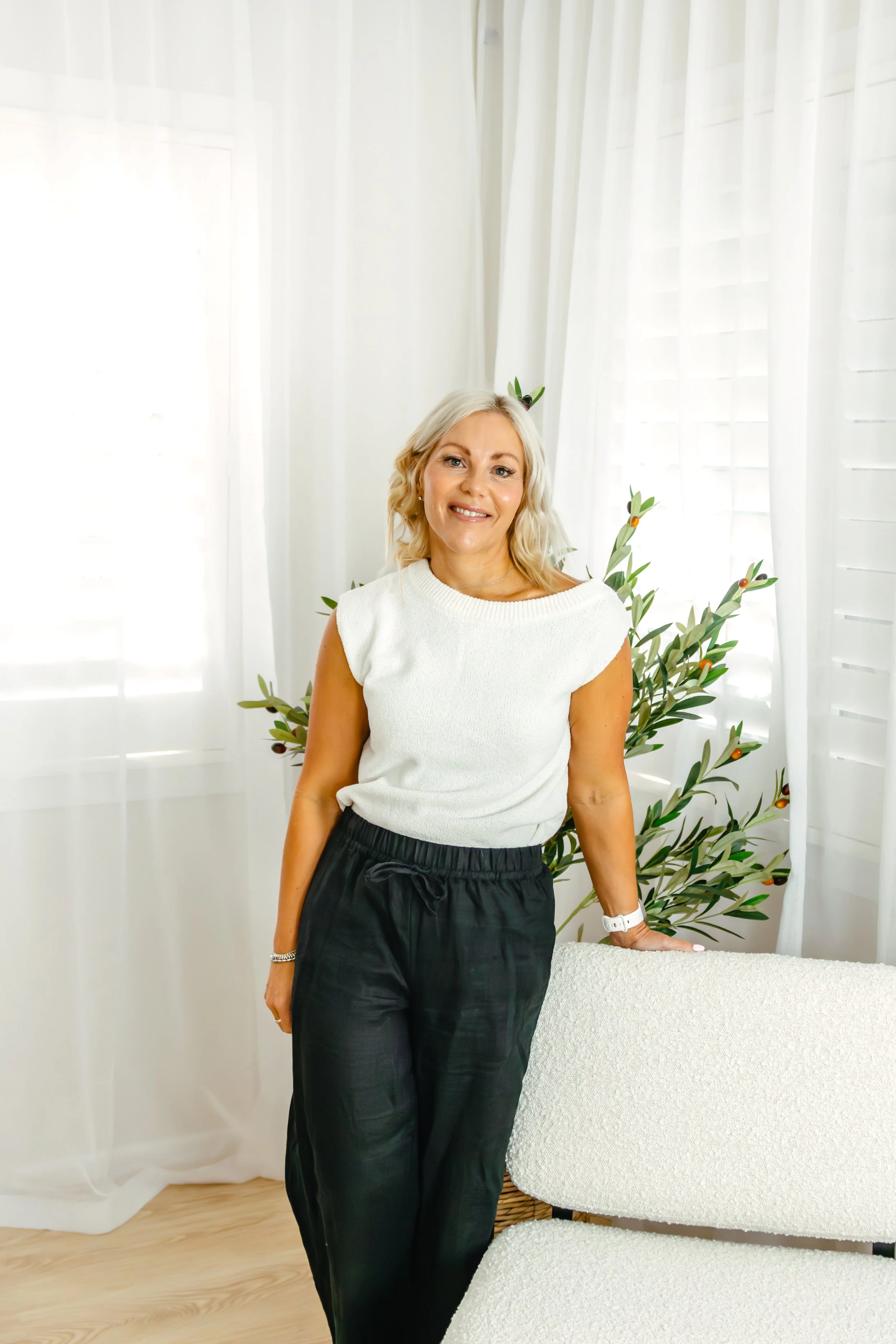 A woman with blonde hair, wearing a white sleeveless top and black pants, stands indoors next to a white textured table with her right hand resting on it. The background includes white curtains, a plant with green leaves and orange berries, and white walls.
