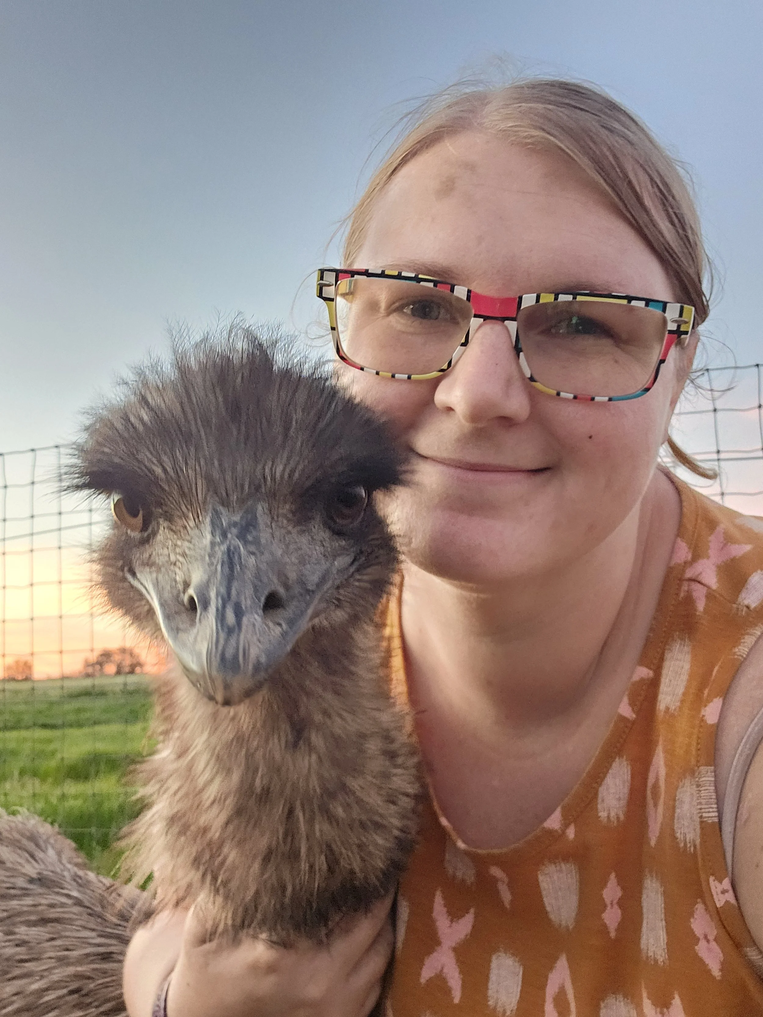 A selfie image of a blonde woman with colorful glasses hugging an emu also looking into the camera with blue skies and fencing behind