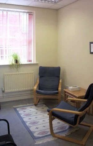 Office corner with two wooden chairs with dark cushions, a wooden side table, a window with pink blinds, a potted plant, and a framed picture on the wall.