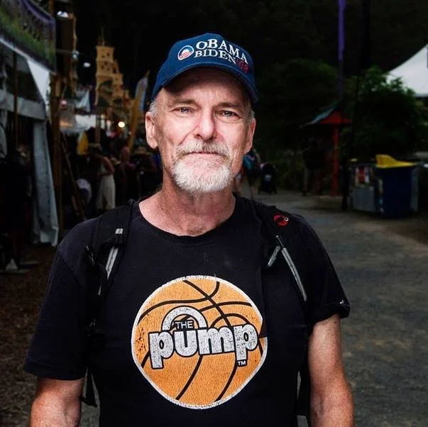 A man with a white beard and mustache wearing a blue 'OBAMA BIDEN' cap and a black t-shirt with a basketball logo and the text 'THE PUMP.' He is outdoors, possibly at a market or fair, with tents and people in the background.