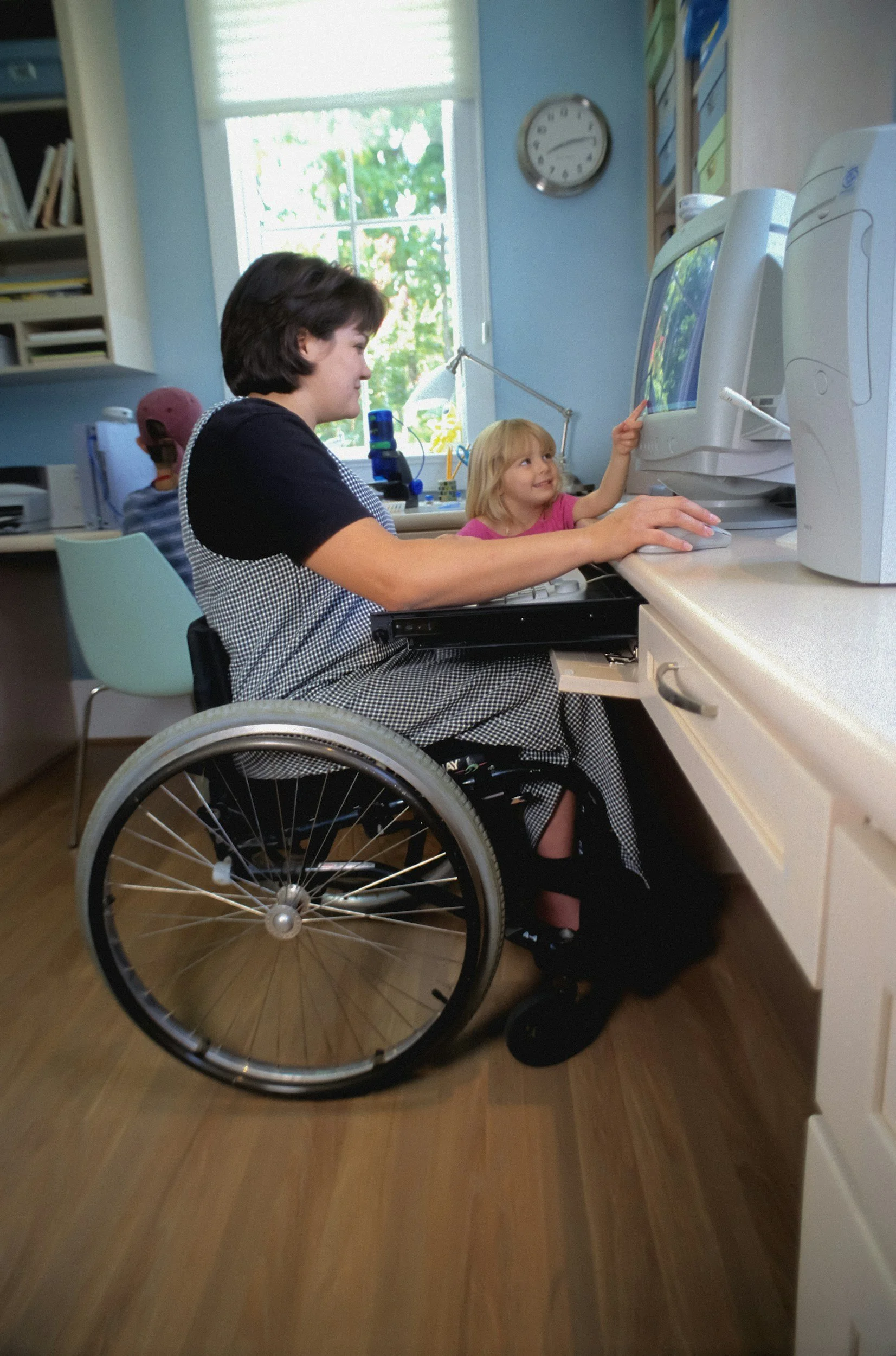 A woman in a wheelchair working on a desktop computer with a young girl pointing at the screen. The scene is set in a room with a window, a clock, and bookshelves.