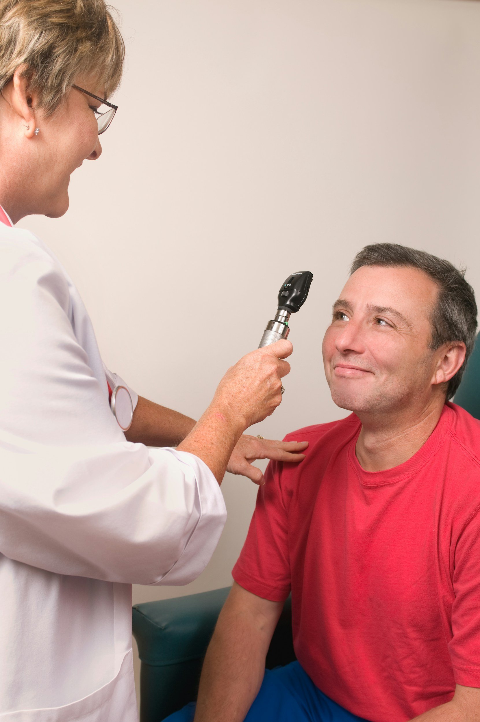 A healthcare professional, wearing glasses and a white coat, is administering a nasal swab test to a man in a red t-shirt and blue pants, who is seated and smiling.