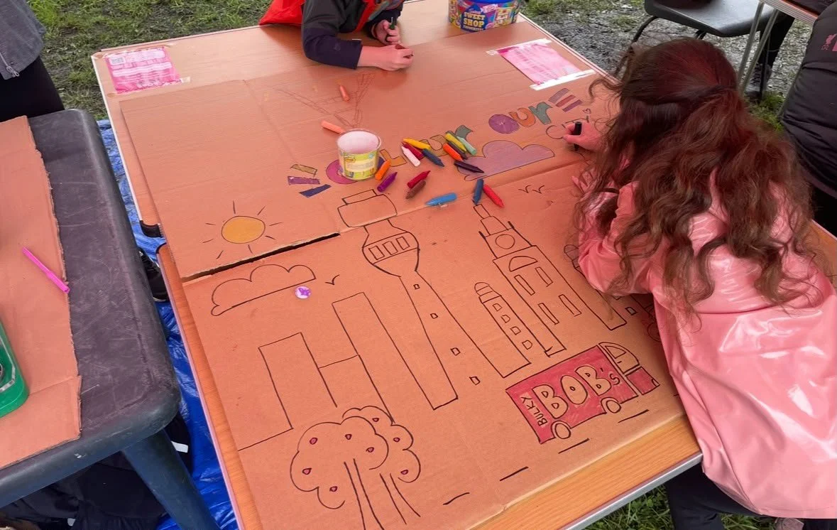 Children leaning on a table colouring in a large carboard picture.