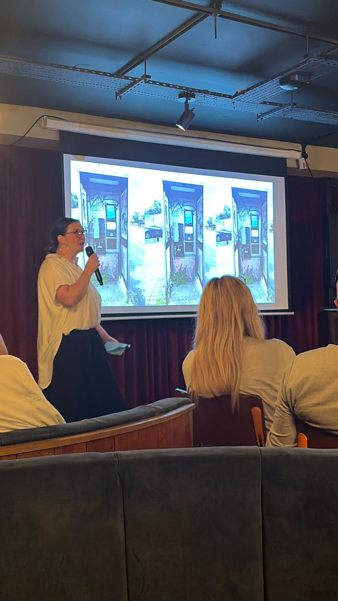 Audience watching a woman in a white top and black skirt, holding a microphone, presenting in front of a large screen.