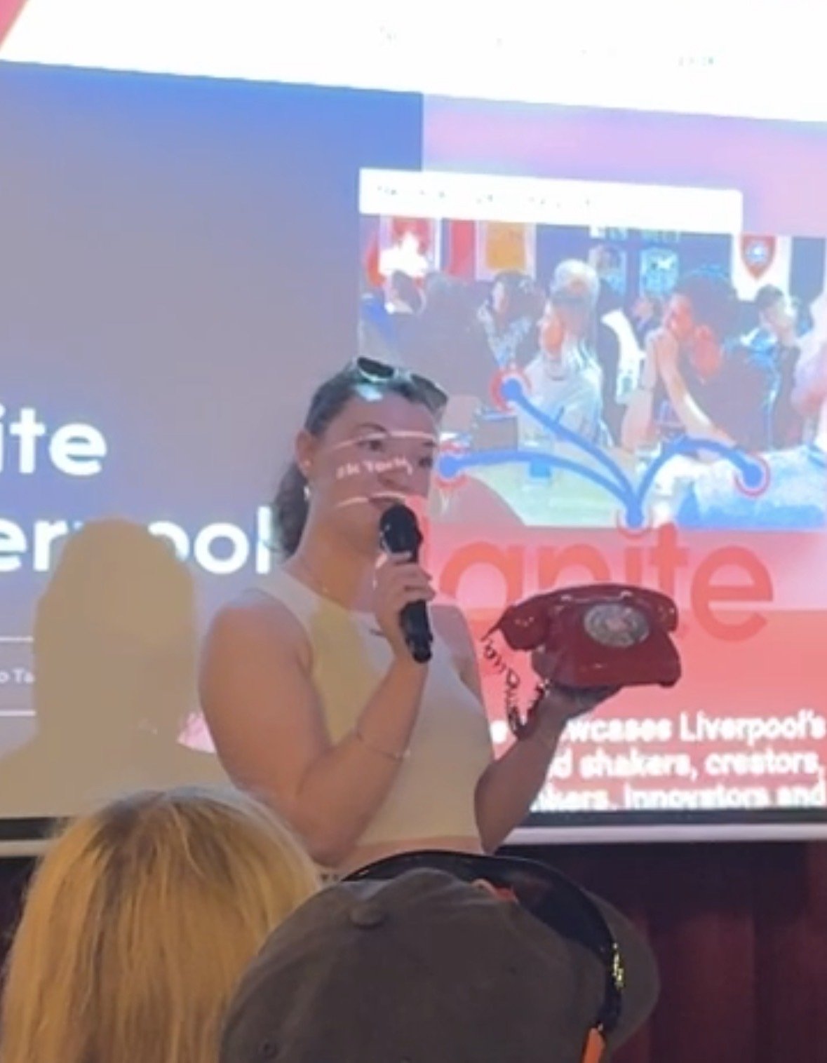 Woman stands in front of large screen holding a red landline phone in her left hand, and a microphone in her right hand. She has brown curly hair and is wearing a white top.