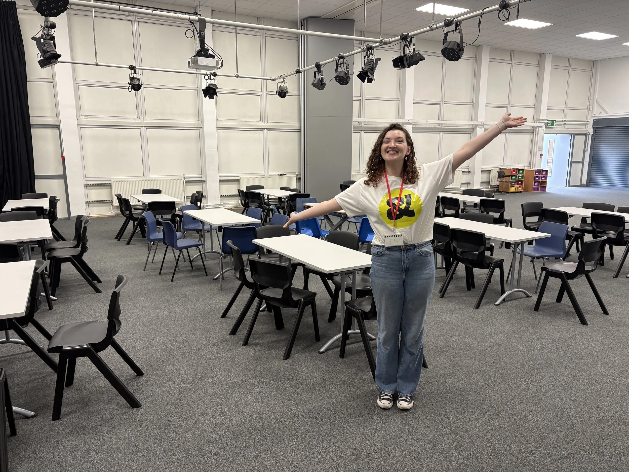 Young woman with brown cursly hair, wearing blue jeans and a white t shirt, stands with her arms out stretched in a large classroom with tables and chairs.
