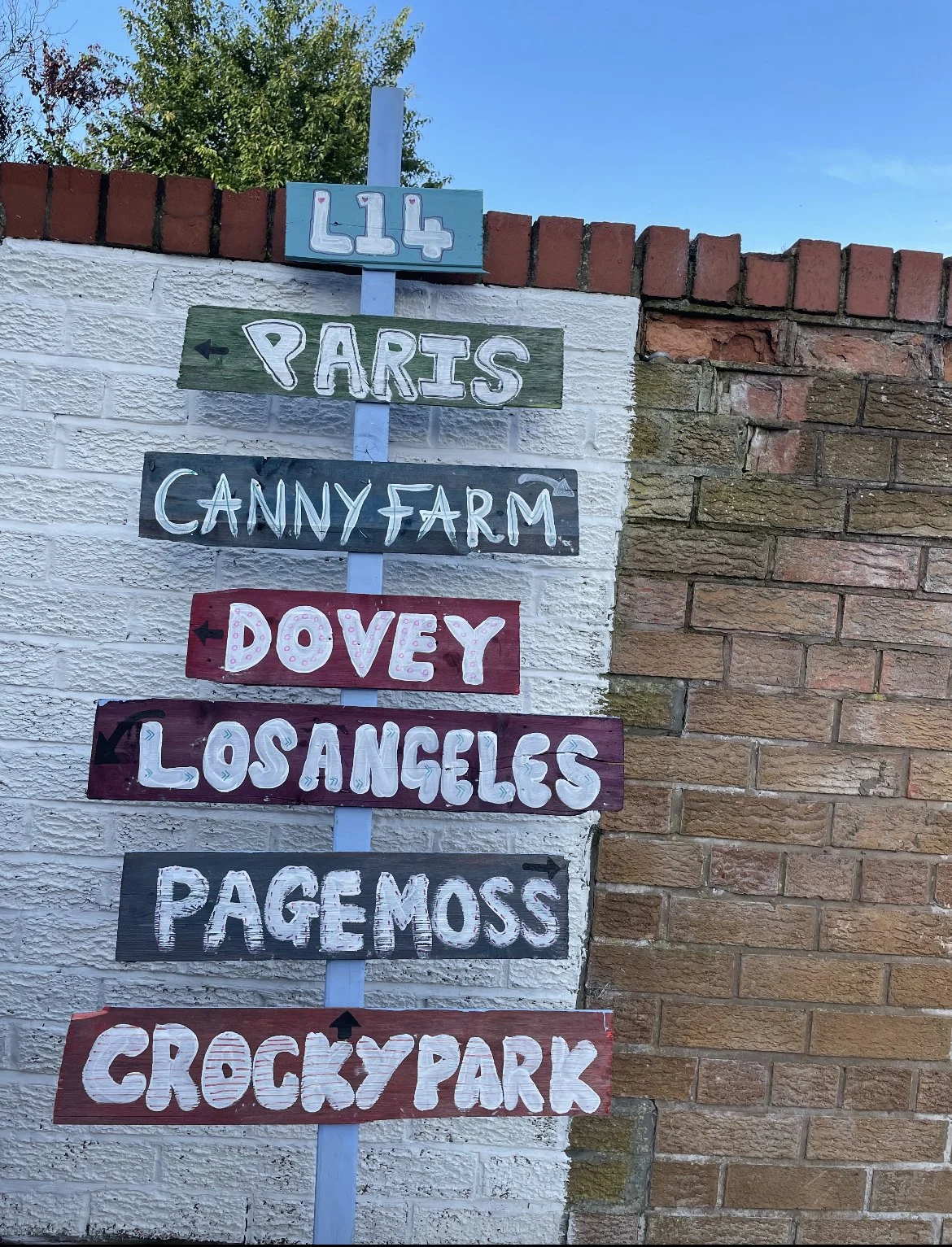 hand painted sign showing global locations and local liverpool suburbs, against a brick wall, blue sky and tree in the background