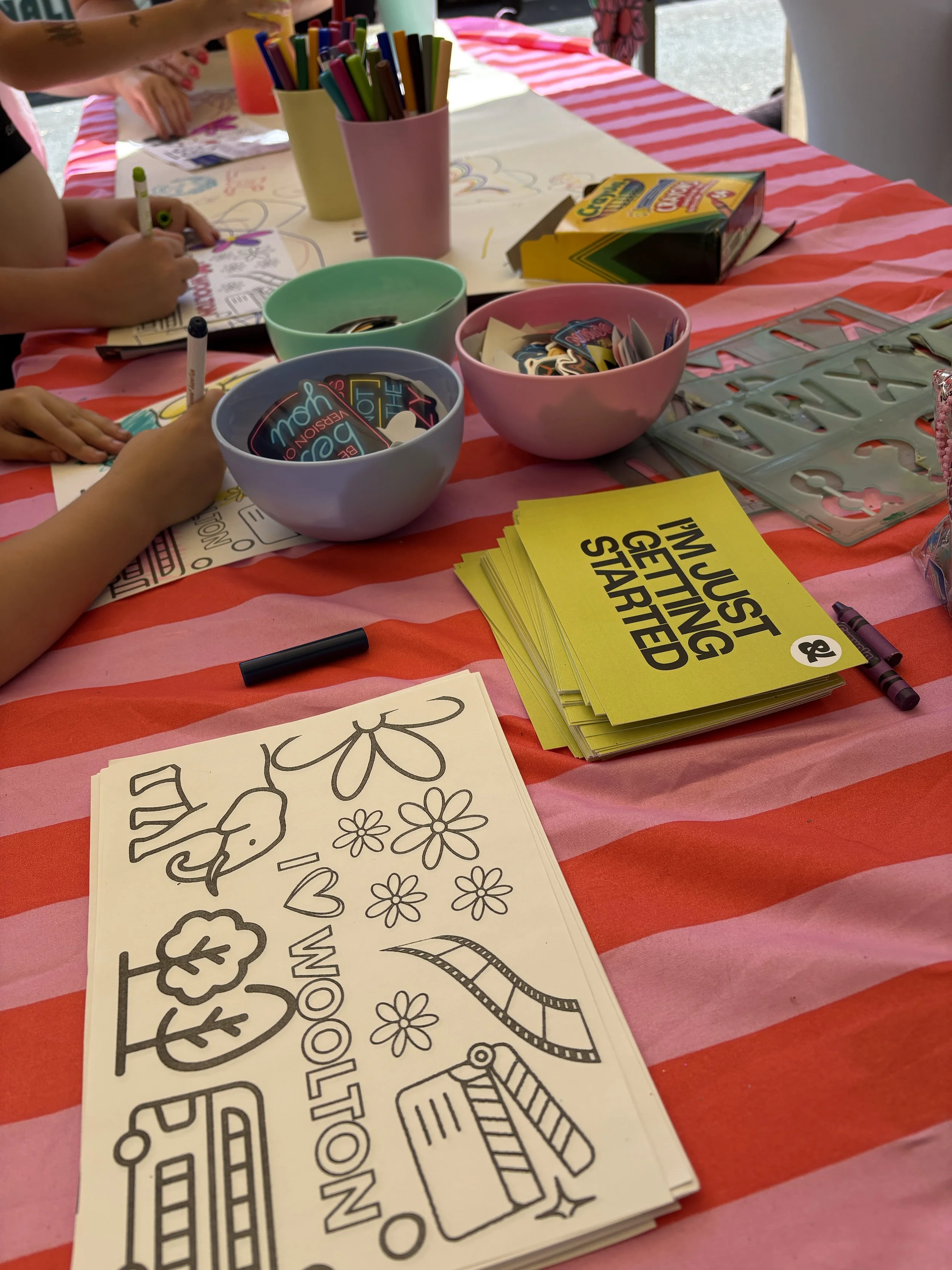 Table top with colouring crayons, stickers in bowls and childrens hands colouring in, on a pink and red stripe tablecloth