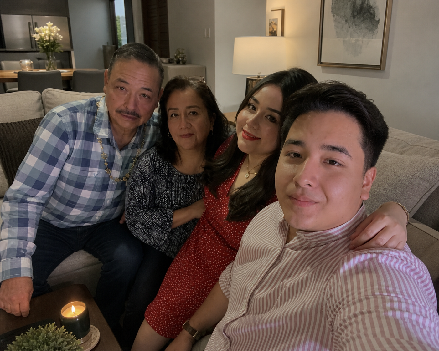 A family of five taking a selfie on a beige couch in a cozy living room, with a candle, plants, and wall art visible.