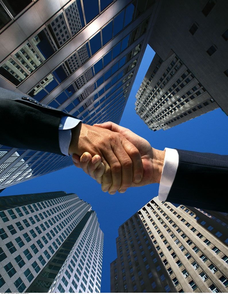 A close-up of two people shaking hands, with tall skyscrapers surrounding them against a clear blue sky.