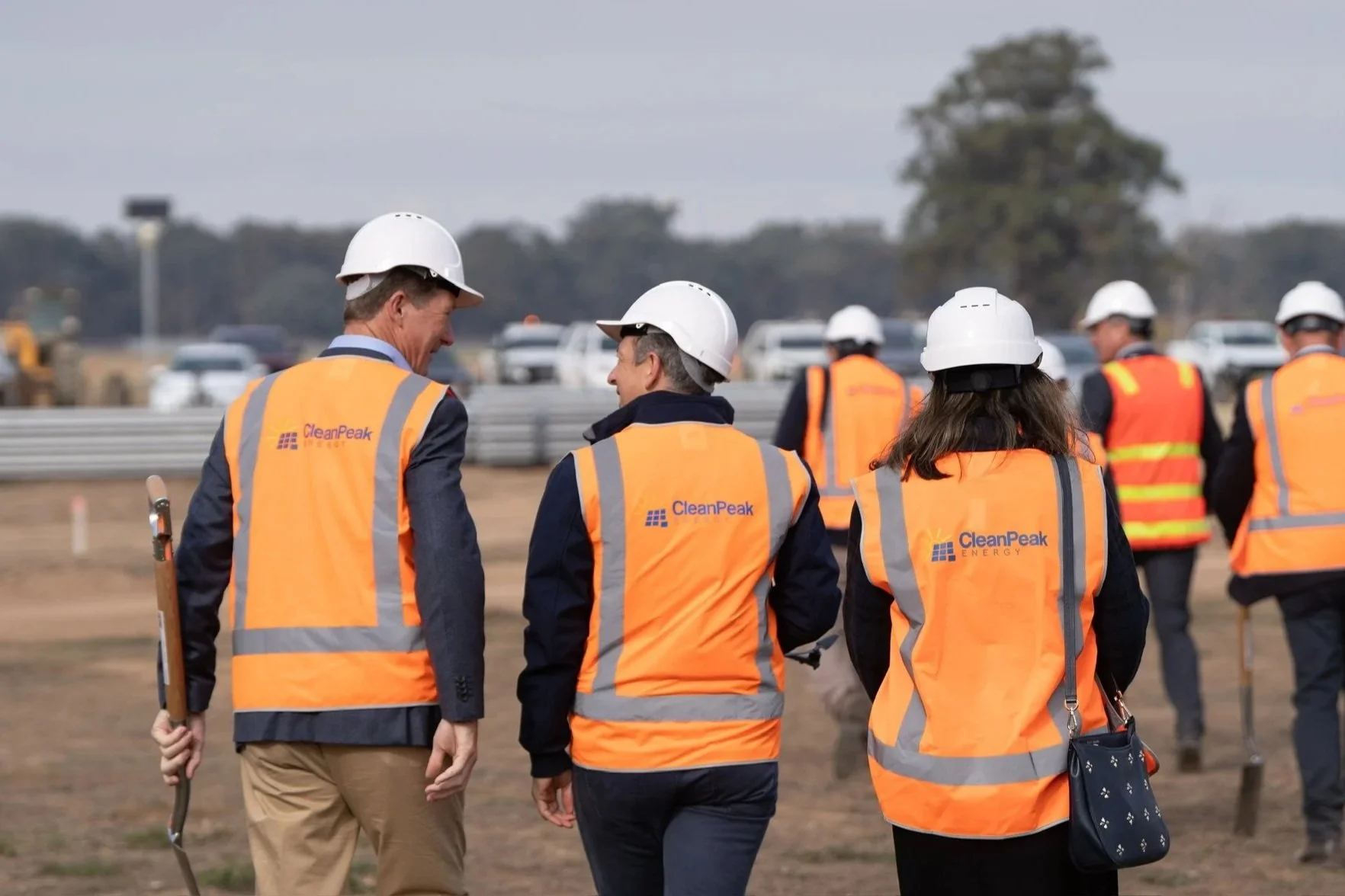 Group of people wearing white hard hats and orange safety vests with 'CleanPeak Energy' logo, walking on a construction site or energy project location.