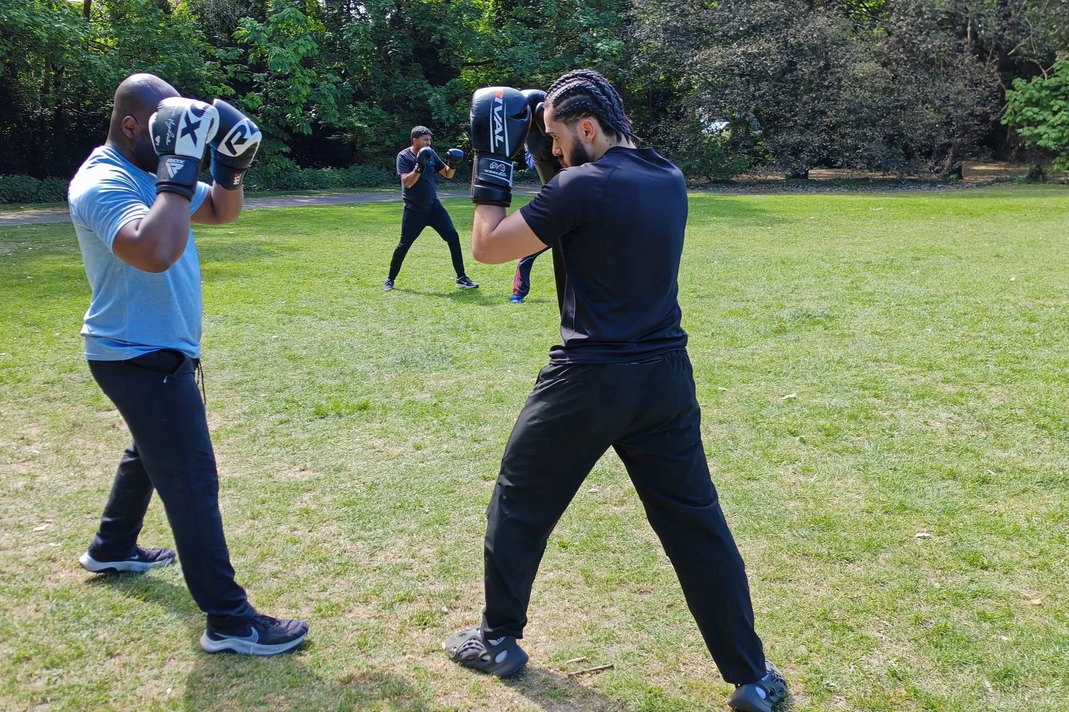 Several people practicing boxing outdoors in a park, with two men in front wearing boxing gloves and pads, and others in the background sparring or training.