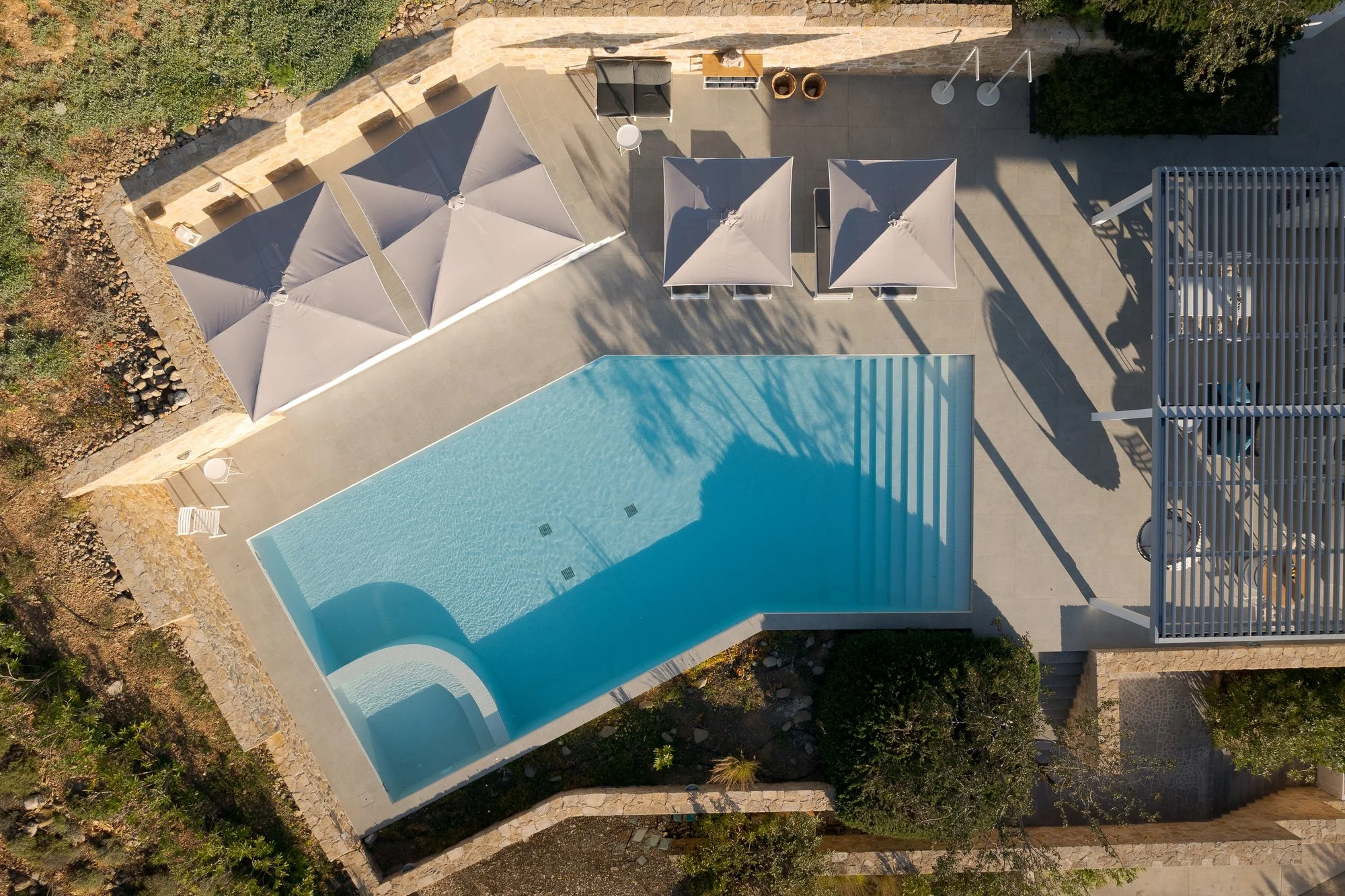 Sunlit pool terrace with umbrellas, lounge chairs, and a slide, surrounded by plants and stone accents at an adults-only stay in South Crete.