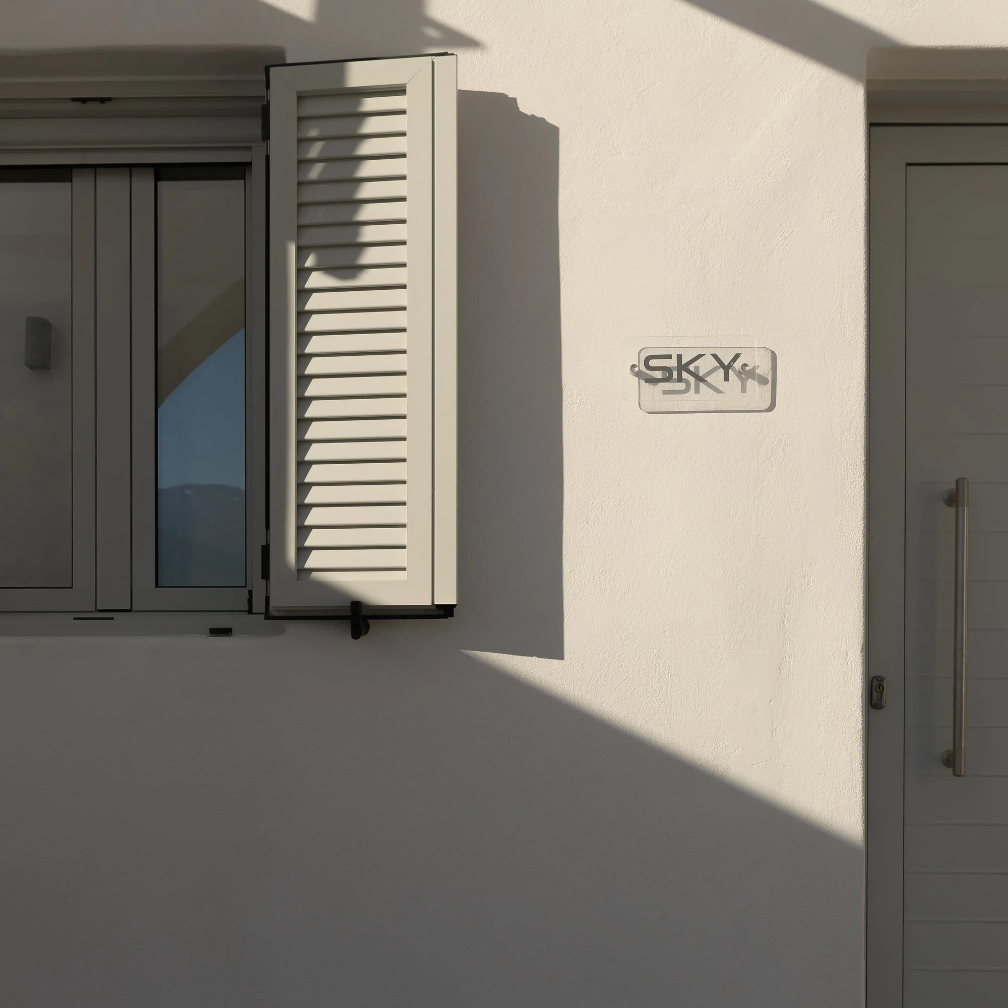 A minimalist white building exterior at Galini Breeze features a white window with louvers and a metallic sign that reads 'SKY' mounted on the wall, with shadows cast across the scene.