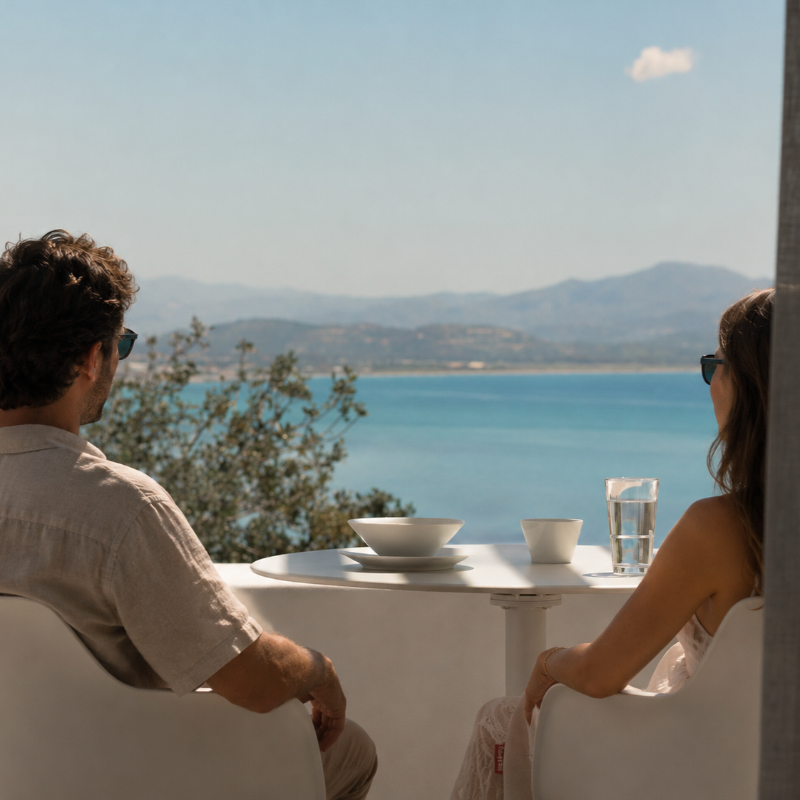 A man and woman sitting at a table on the private terrace at an adults-only resort in South Crete with dishes and a glass of water, overlooking a lake and mountains.