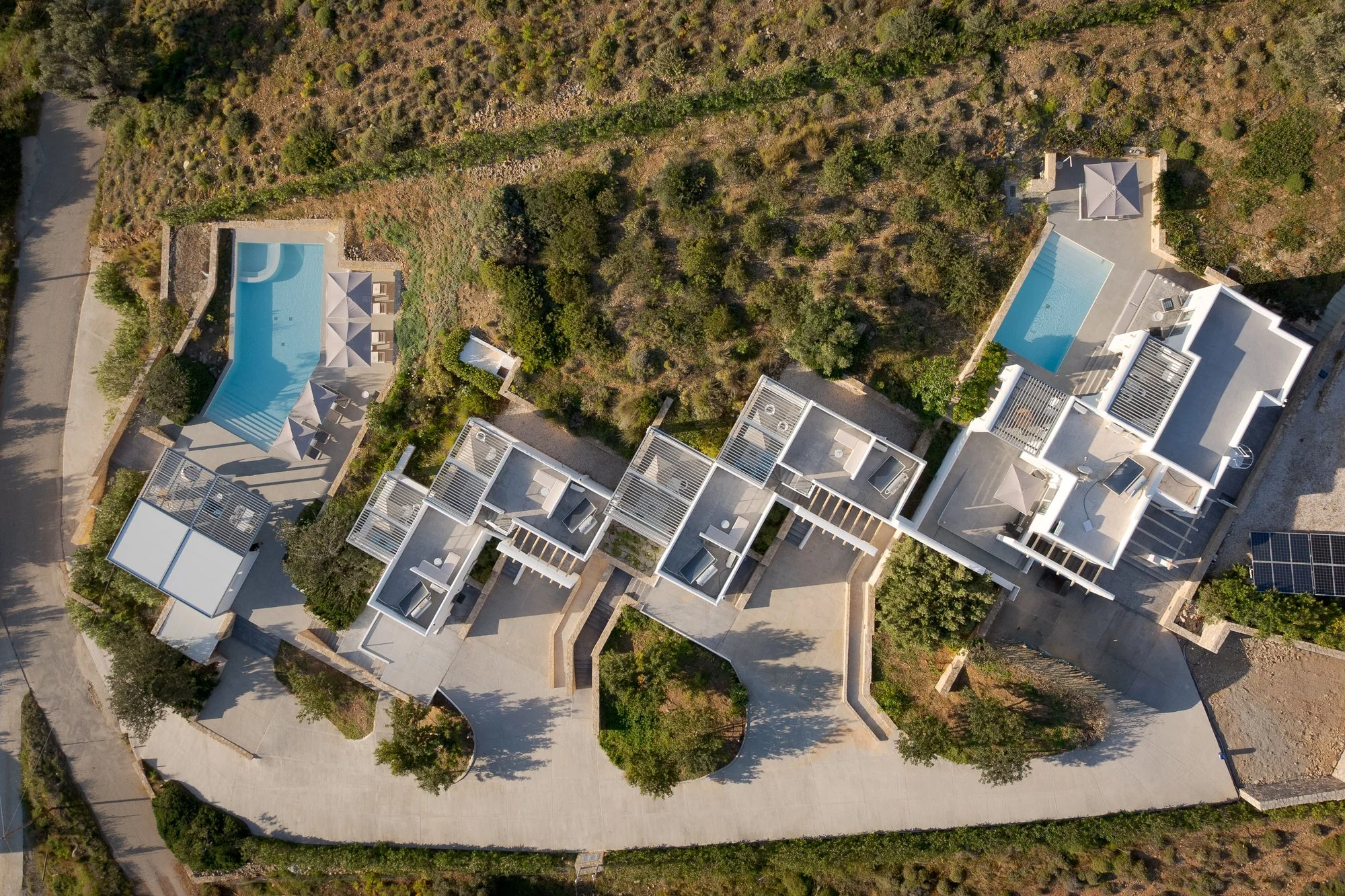 An aerial view the Galini Breeze hillside resort with heated swimming pools and modern white buildings, surrounded by greenery and trees.