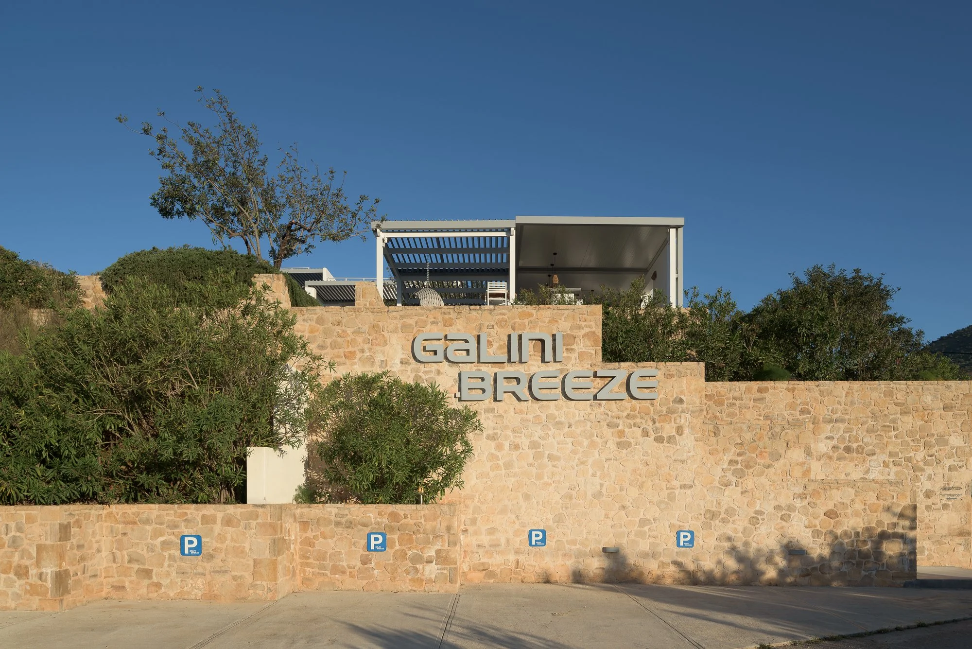 A modern building with a rooftop terrace and a large sign that reads 'GALINI BREEZE' on a stone wall beneath the terrace. The area in front has parking spots. The sky is clear and blue, with some greenery around.