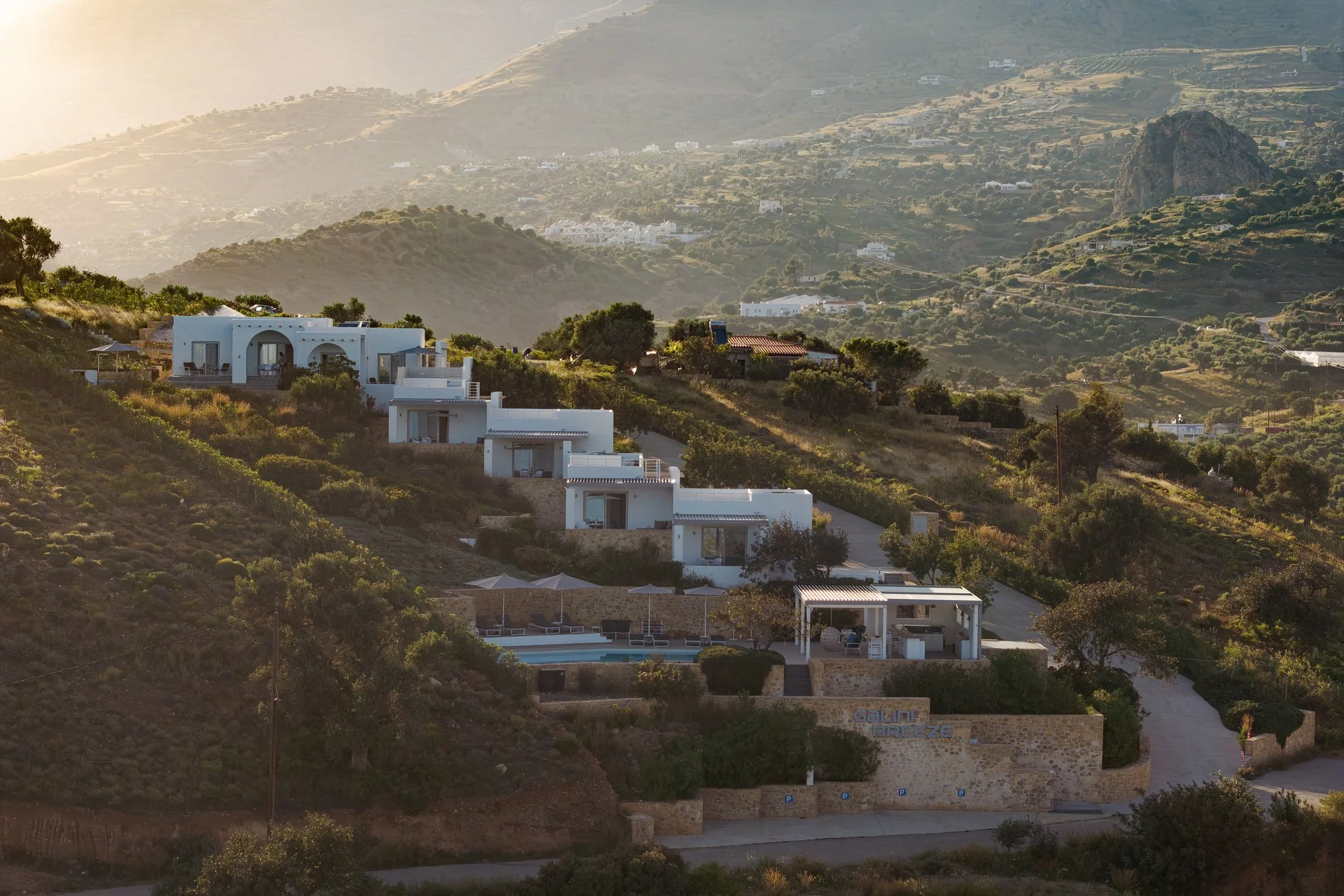White hillside studios surrounded by greenery, overlooking a distant mountain range in South Crete, at sunset.