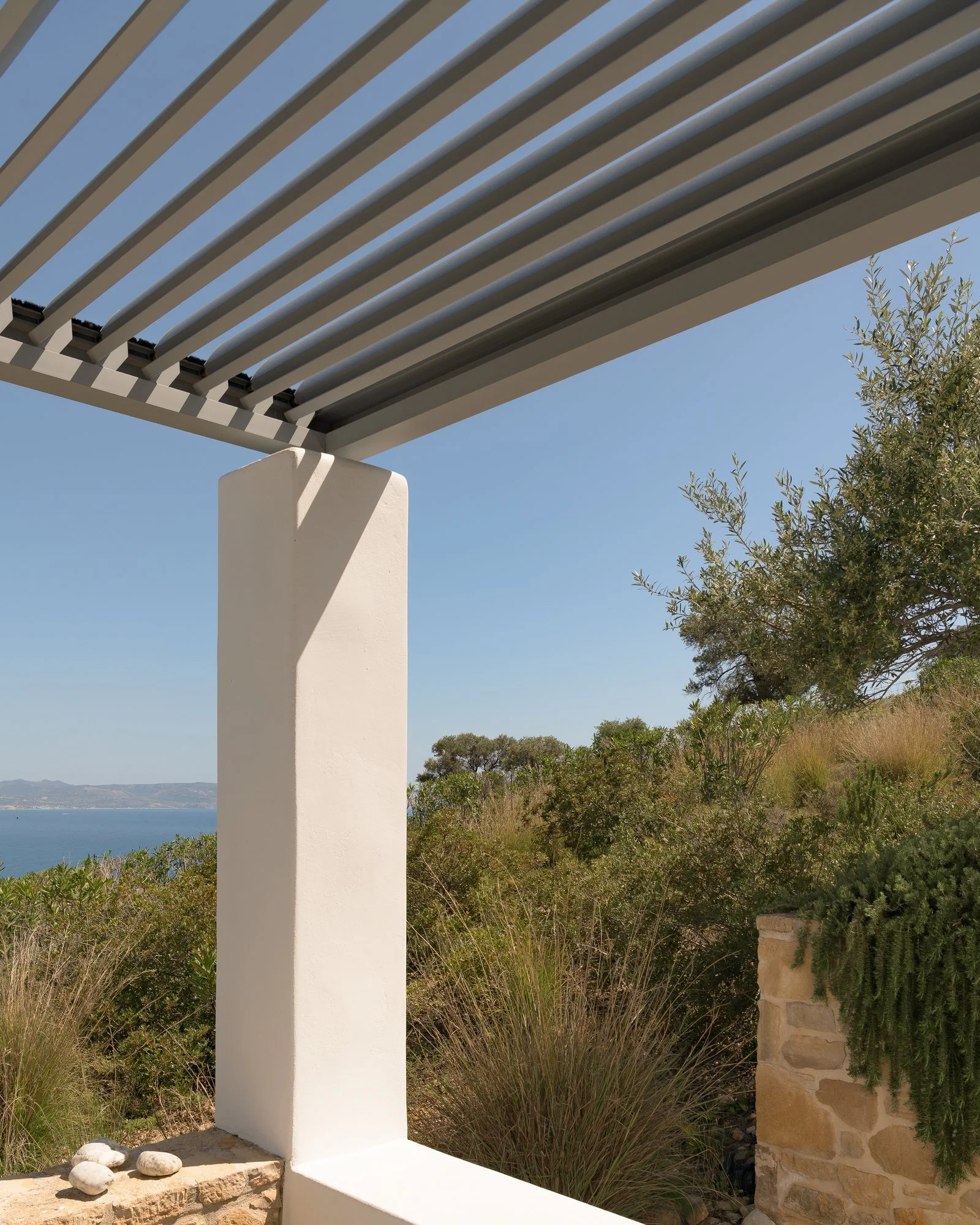 White architectural structure with rounded stones at its base, situated in a natural landscape with bushes, trees, and a view of the water and distant land under a clear blue sky.