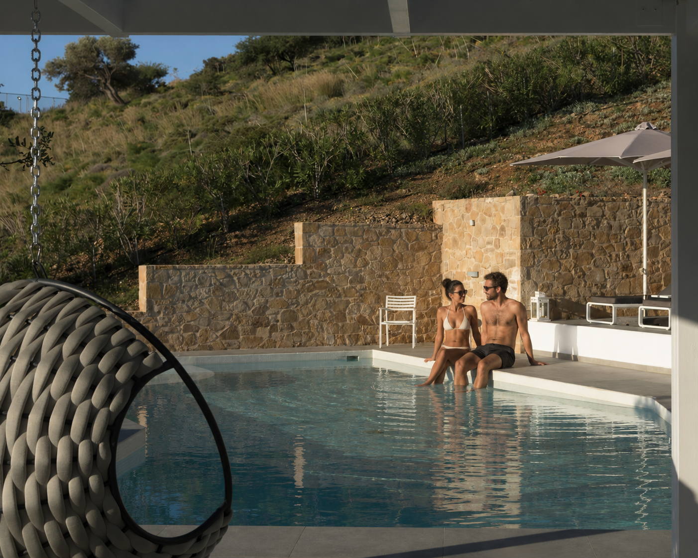 A couple sitting on the edge of the heated swimming pool at Galini Breeze, Agia Galini, with the woman wearing a bikini and the man in swim trunks, overlooking a hillside with greenery and a stone wall, during a sunny day.