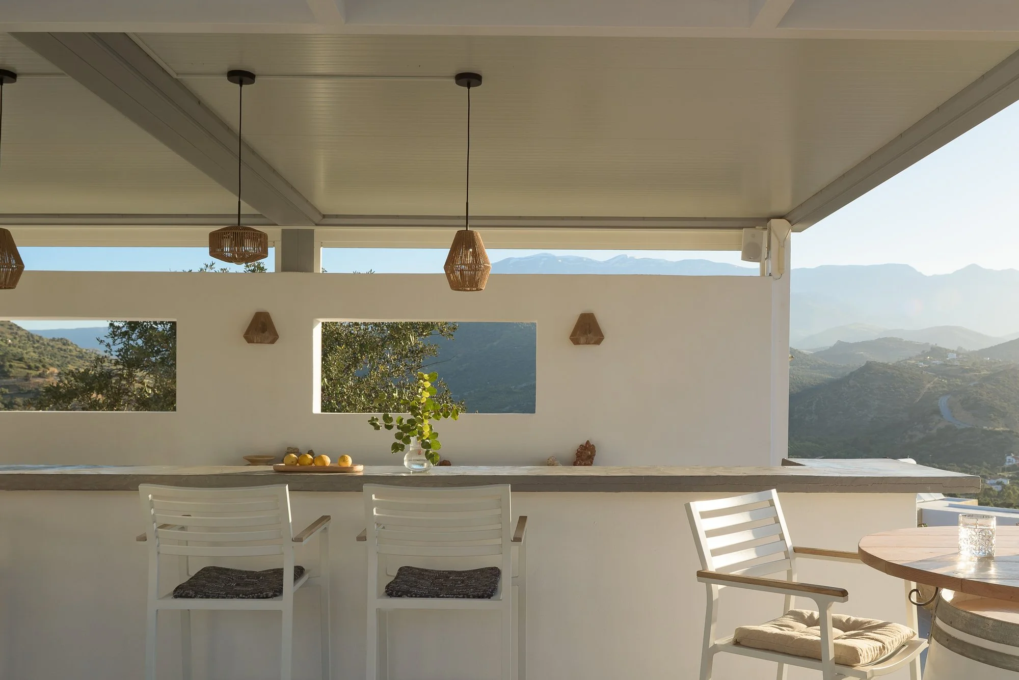 A modern outdoor patio area with white chairs, a table, and hanging wicker pendant lights. Mountains and blue sky can be seen in the background through large rectangular openings in the wall at Galini Breeze, Agia Galini.