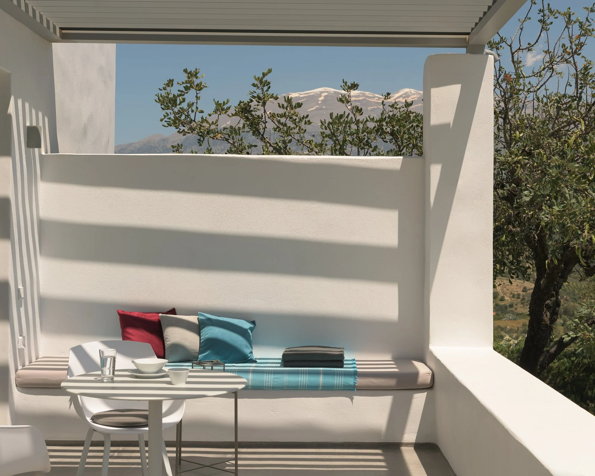 Outdoor balcony at a studio at Galini Breeze with white walls and modern furniture, including a table, chairs, colorful pillows, and a built-in bench with cushions, overlooking green trees and mountains in the background.