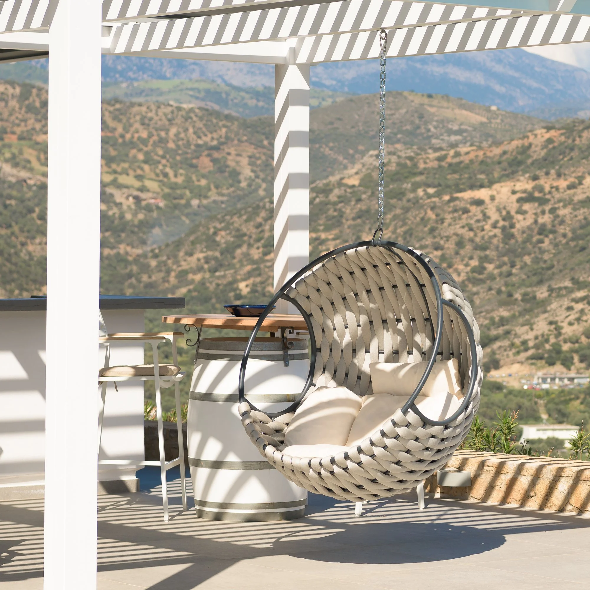 Outdoor lounge terrace with woven hanging chair and white pergola casting shadows, South Cretan mountain landscape in the background.