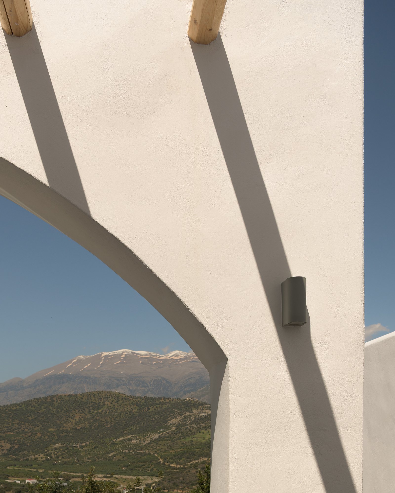 Close-up of a modern white architectural structure with curved and straight elements, shadows cast by supports, with mountains and blue sky in the background.