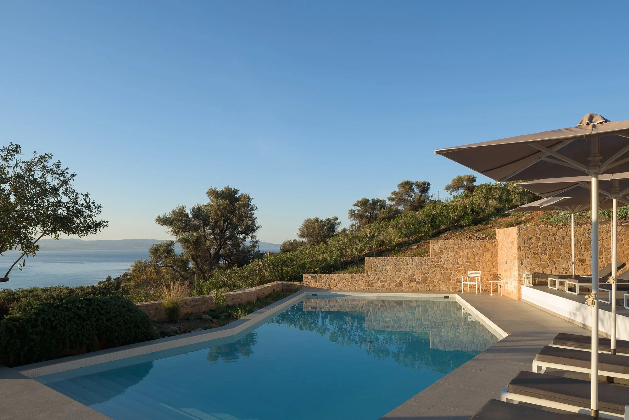A modern swimming pool with a view of the ocean, surrounded by lounge chairs with umbrellas, trees, and a clear blue sky.