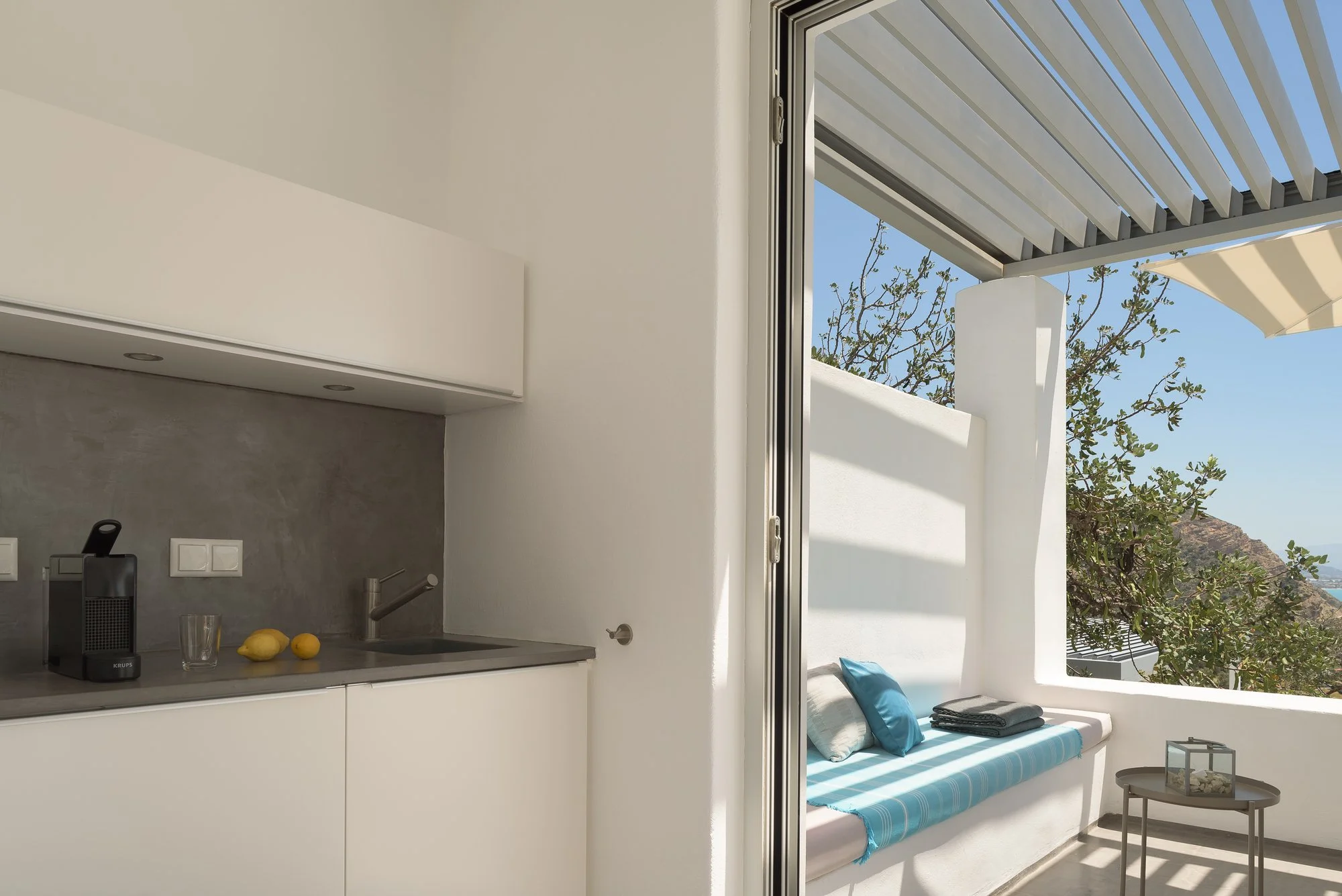 Minimalist kitchen and shaded terrace with outdoor seating at Galini Breeze in Agia Galini. The room is opening to a balcony with outdoor seating, blue cushions, a white wall, and trees visible outside.