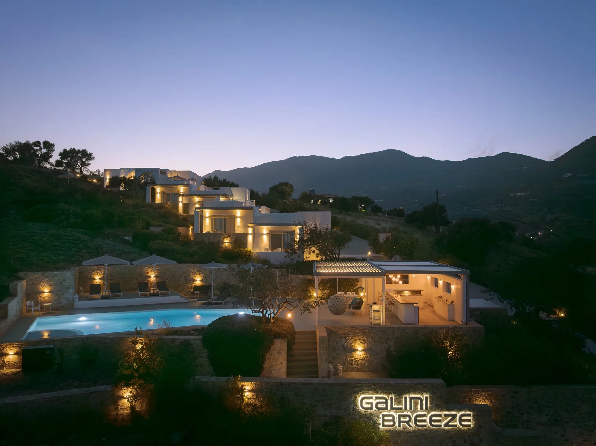 Luxury modern house with illuminated exterior, swimming pool, and scenic mountain view at dusk, with illuminated 'Galini Breeze' sign.