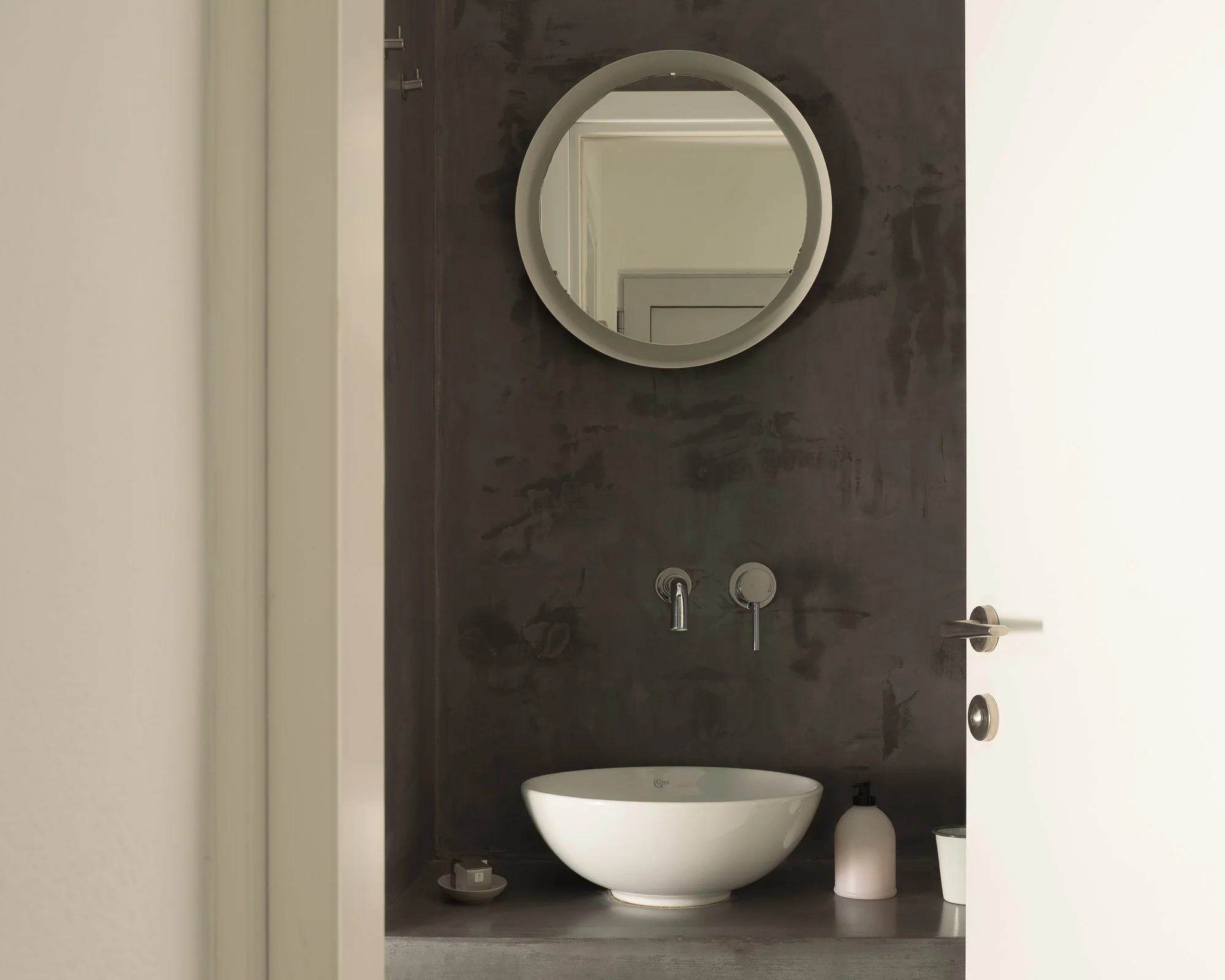 A modern bathroom with a dark wall, a round wall mirror, a white toilet, and bathroom accessories including a soap dispenser and a small container.