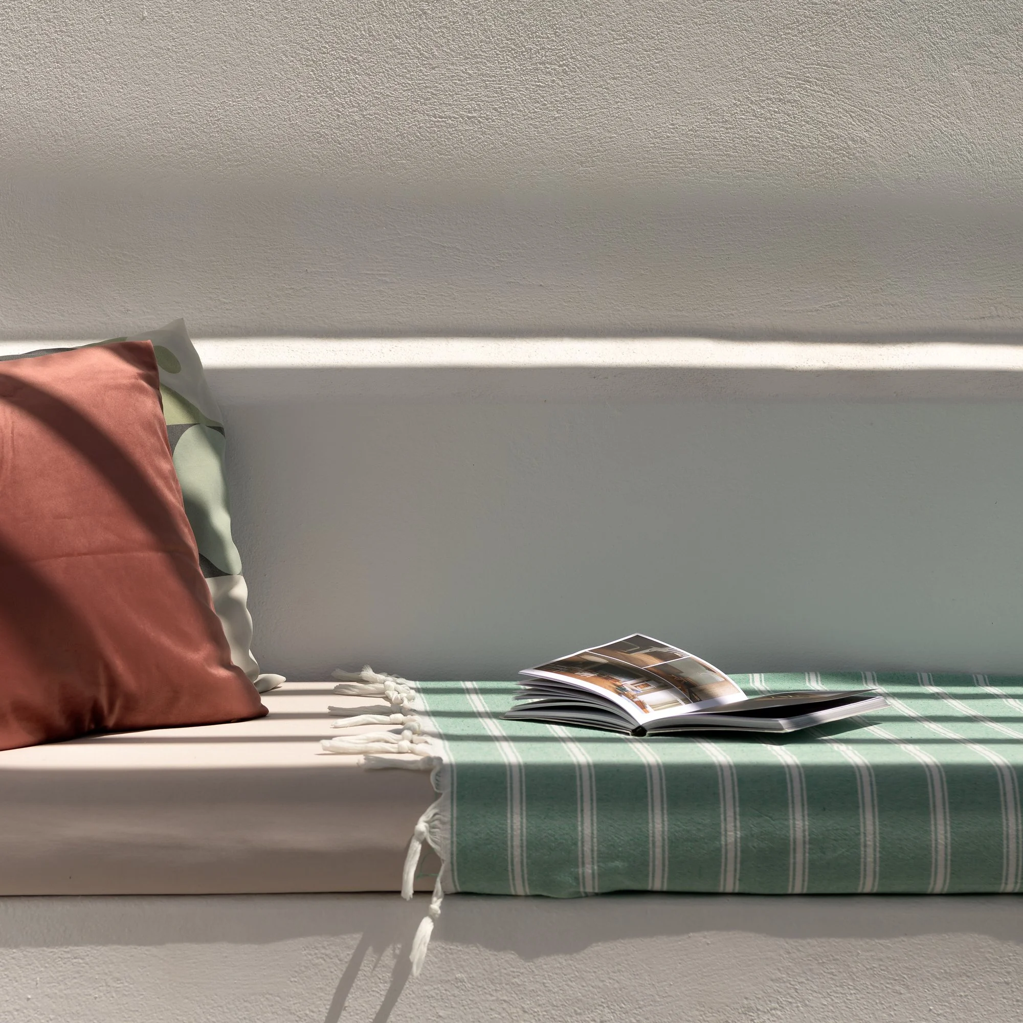 Close-up of a bed with pillows, a green and white checkered blanket, and an open magazine, against a textured wall with sunlight casting shadows.