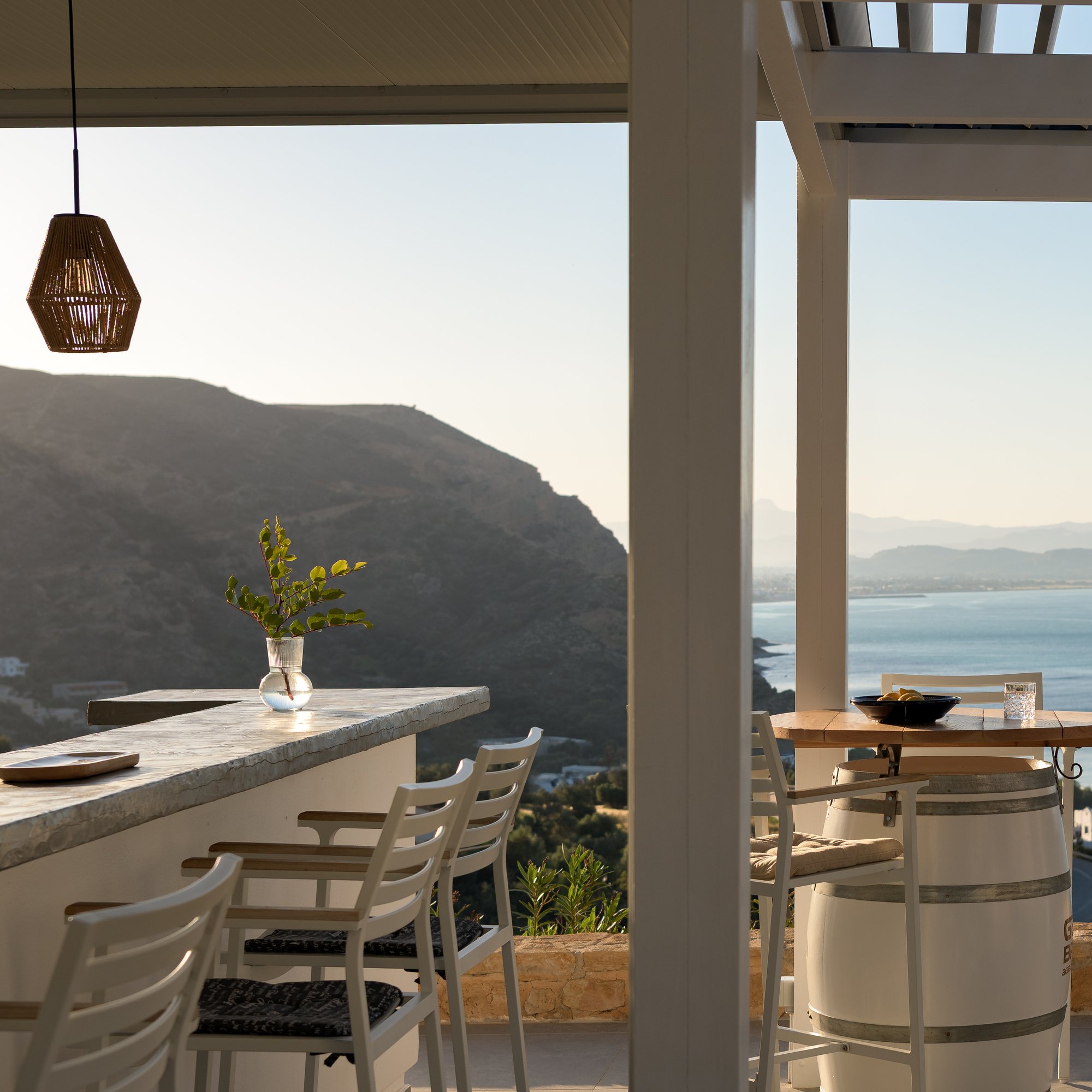 Minimalist pool bar and terrace with sea views at Galini Breeze, with white chairs, a marble counter, decorative items including a glass cup, a bowl of lemons, and a vase with greenery, and wooden beams with natural light coming through.